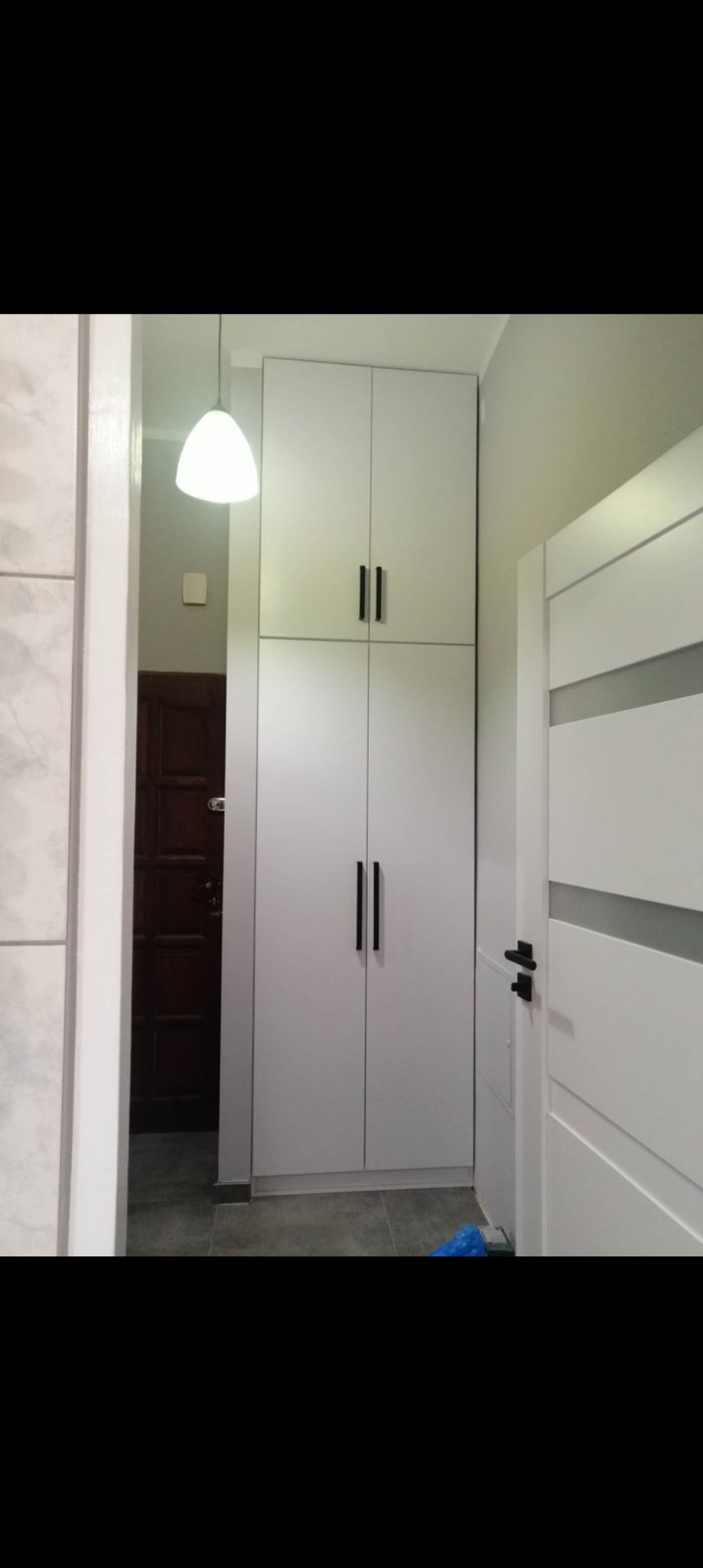 Biała szafa do sufitu z czarnymi, podłużnymi uchwytami, ustawiona w przedpokoju obok białych drzwi z szarym paskiem, oświetlona wiszącą lampą z białym kloszem.