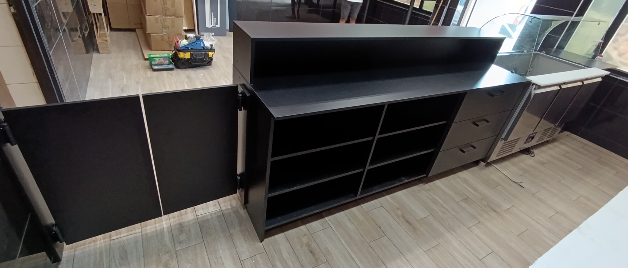 Czarna lada sklepowa z otwartymi drzwiczkami szafki, widoczne półki, obok chłodziarka ekspozycyjna, na podłodze płytki imitujące drewno, w tle narzędzia.