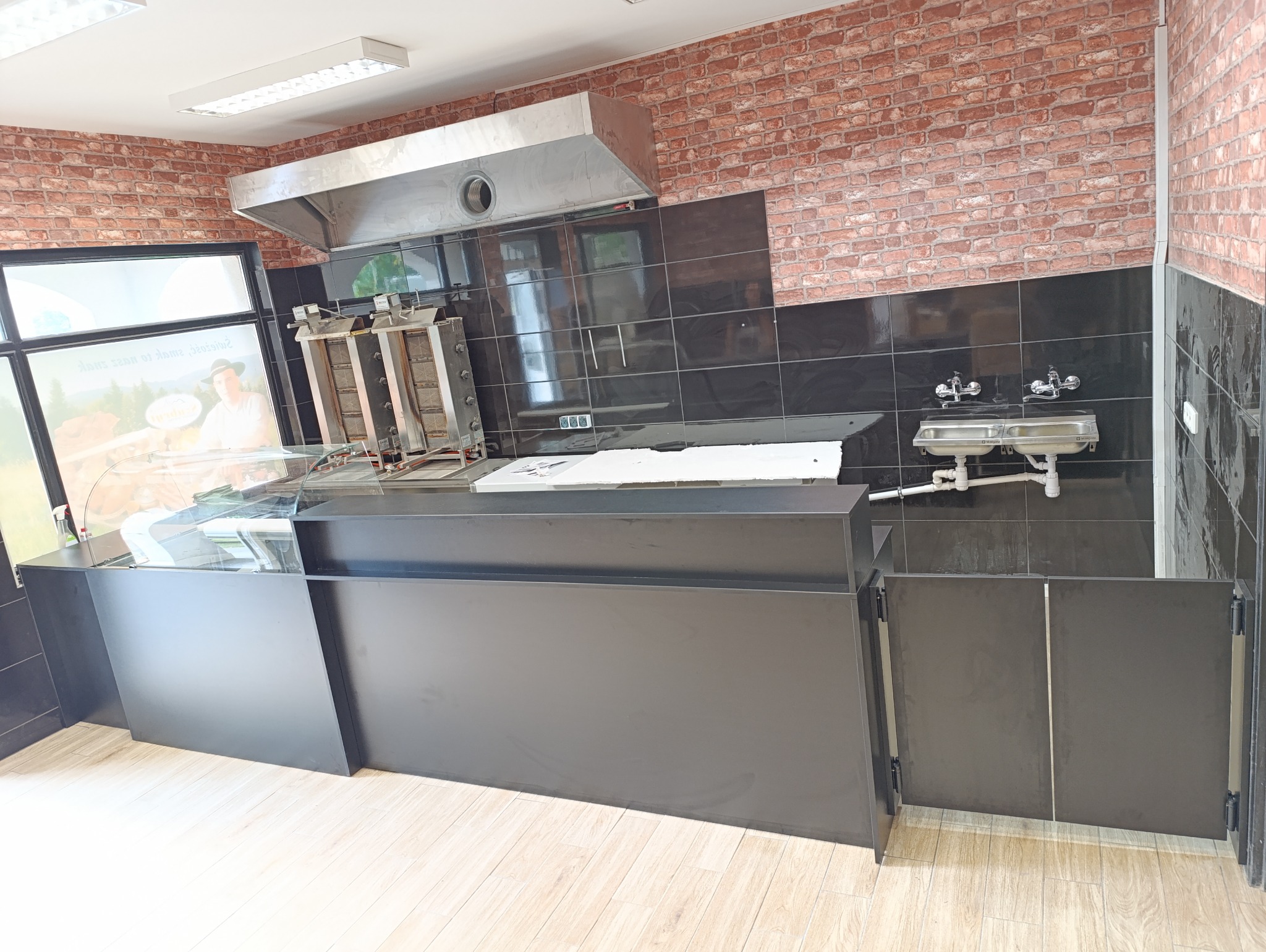 Wnętrze lokalu gastronomicznego z wyposażeniem: lada chłodnicza z szybą, opiekacz do mięsa, okap, umywalka i ściany wyłożone czarnymi płytkami oraz cegłą.