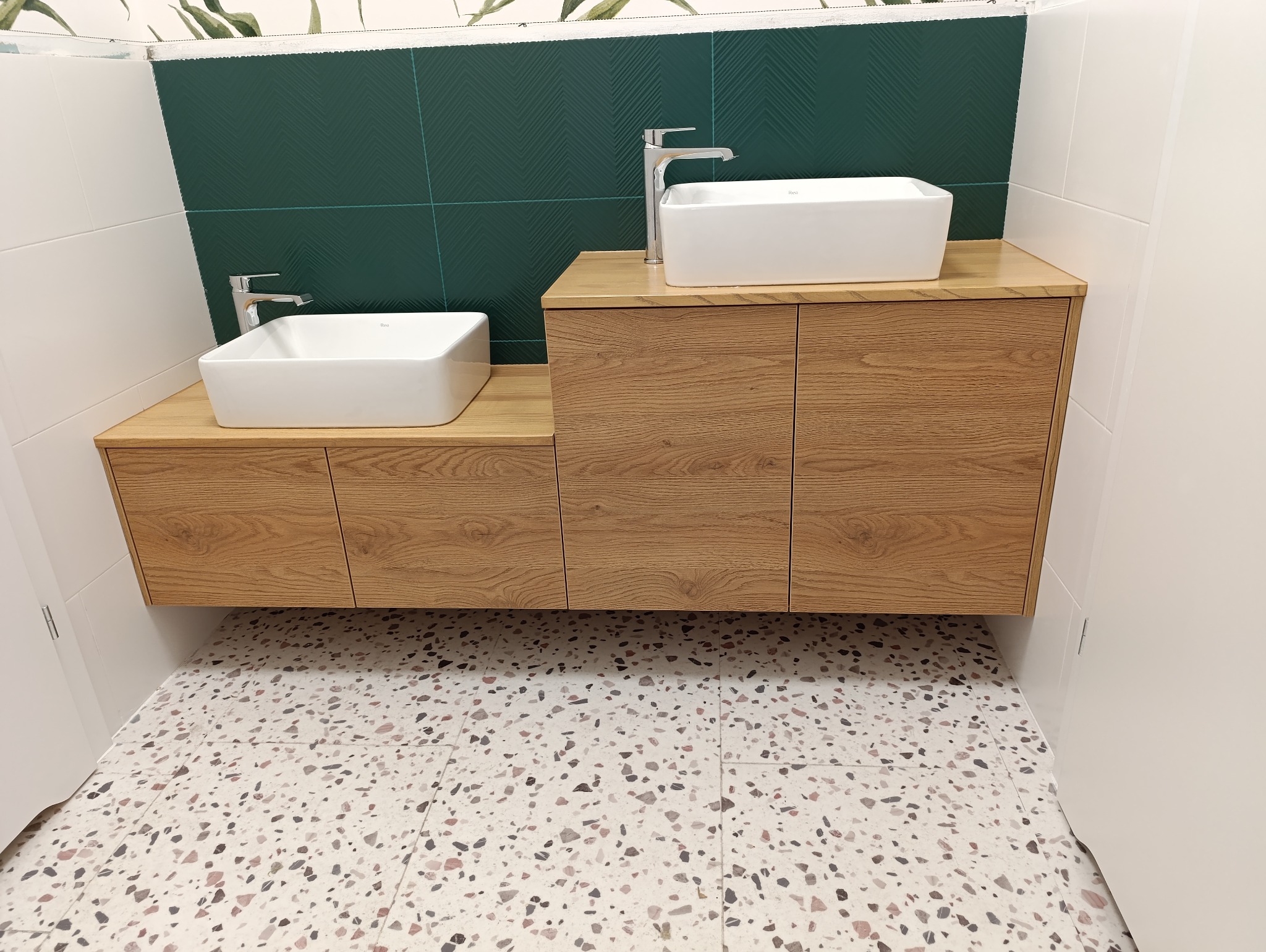 Dwie umywalki nablatowe o kwadratowym kształcie, zamontowane na drewnianych szafkach w łazience z zielonymi i białymi płytkami oraz podłogą w stylu lastryko.