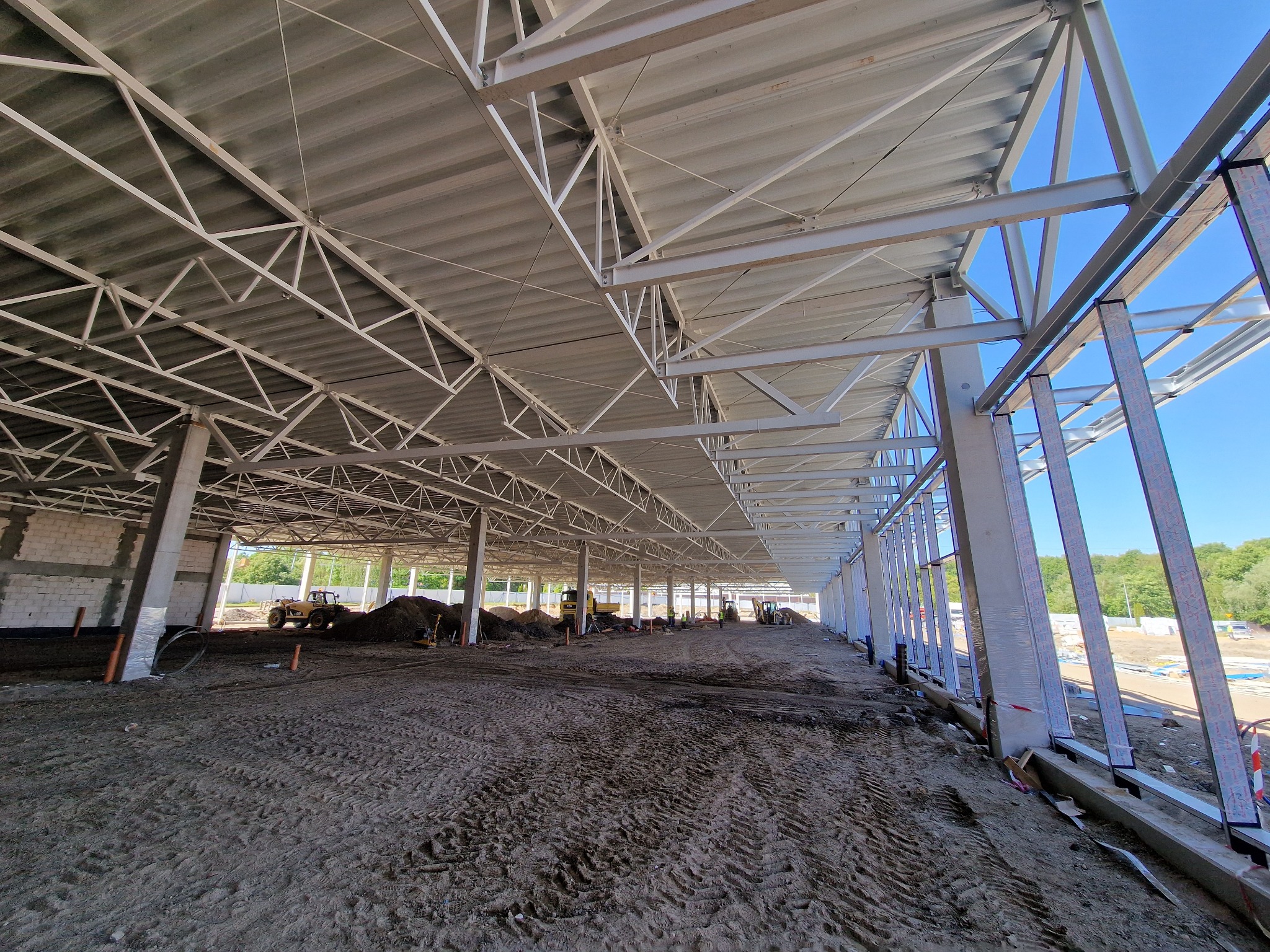Wnętrze hali w budowie w Bydgoszczy, widoczne stalowe konstrukcje nośne dachu i ścian, ziemia na podłożu oraz maszyny budowlane w oddali.