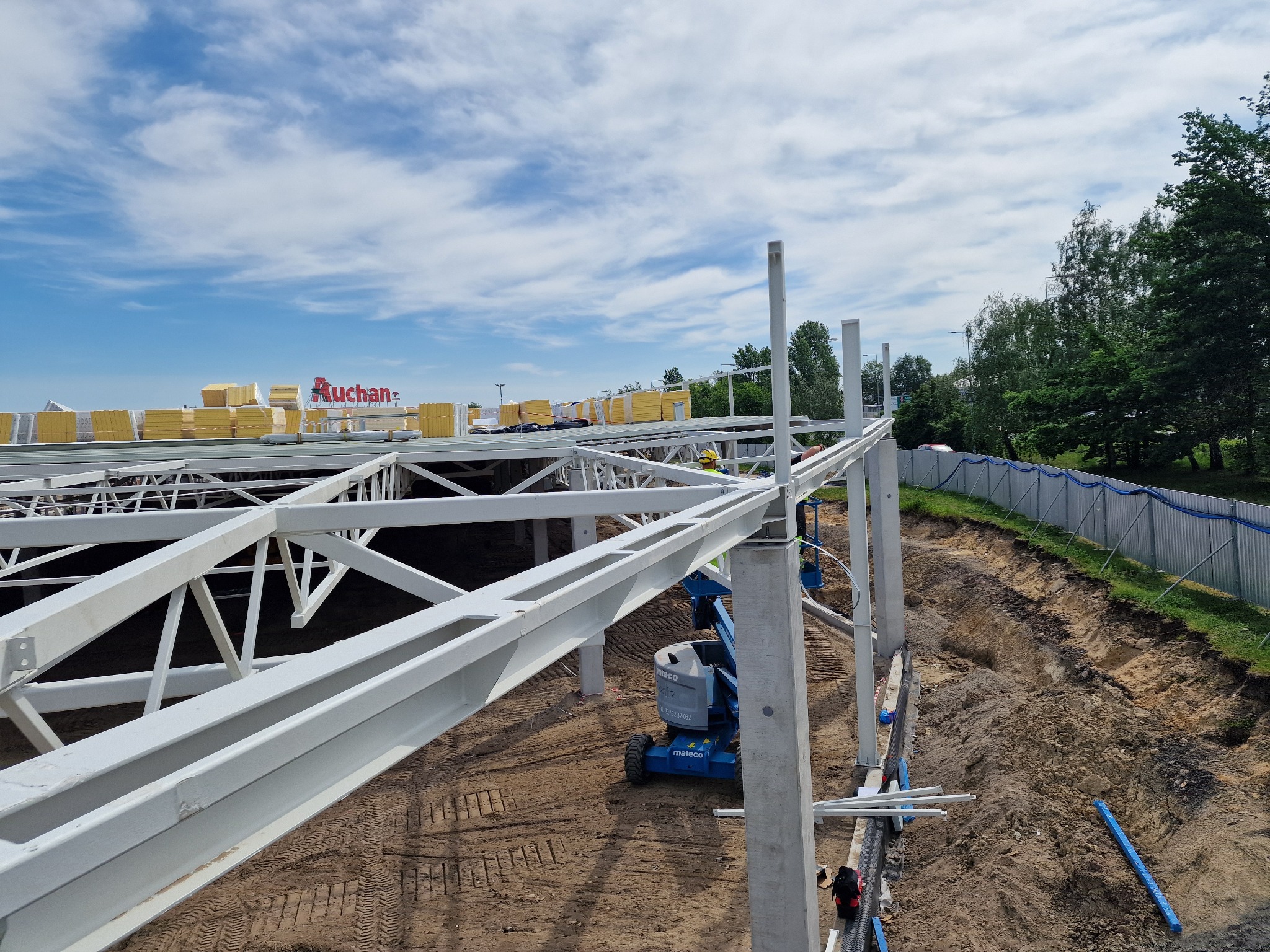 Wysoka perspektywa na budowę hali stalowej z widocznym niebieskim podnośnikiem Mateco, elementami konstrukcyjnymi w kolorze białym i stertą płyt na dachu, w tle logo Auchan.