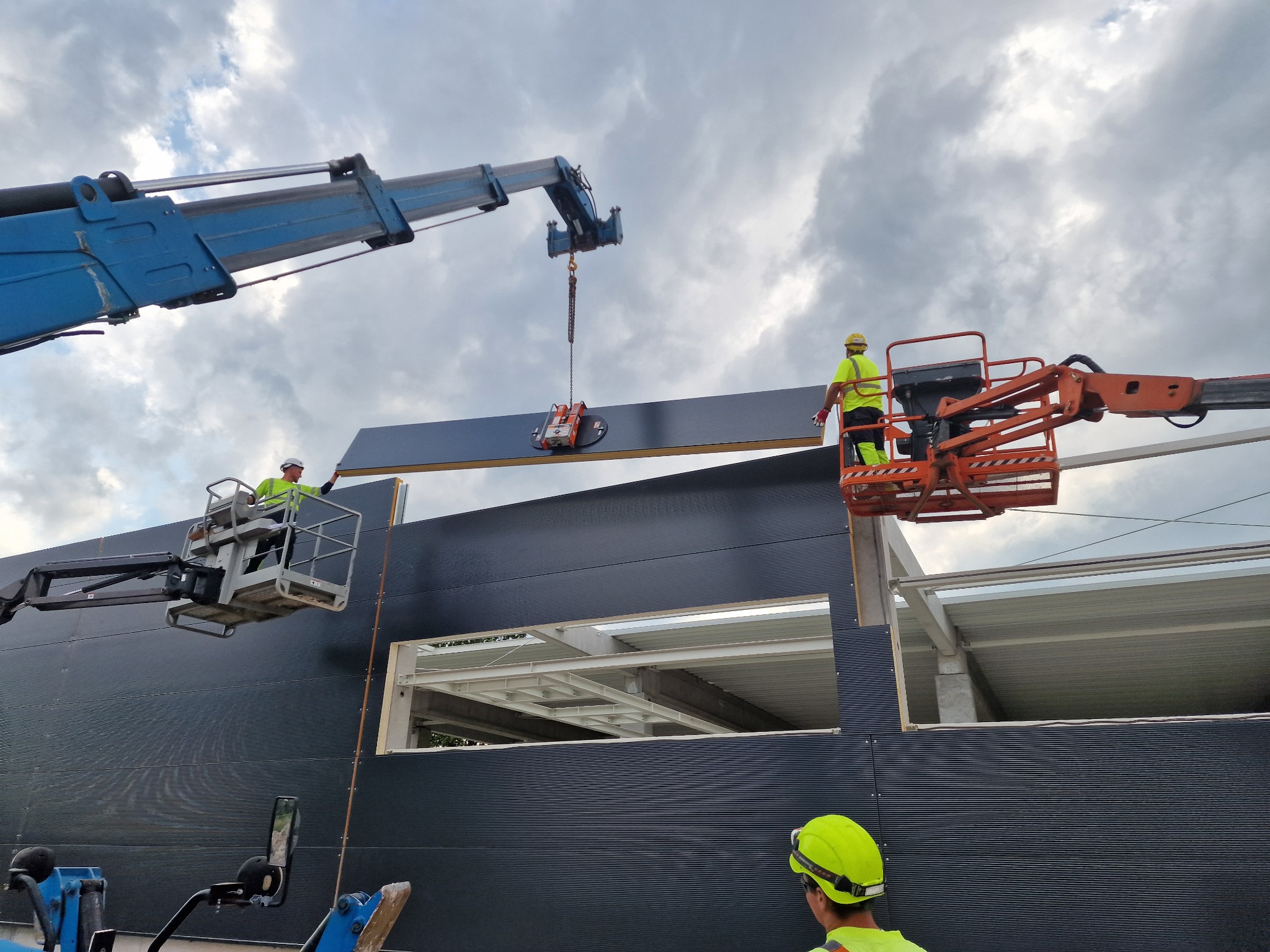 Montaż ciemnej płyty warstwowej na konstrukcji stalowej hali za pomocą przyssawki podwieszonej do dźwigu, z udziałem dwóch podnośników koszowych i pracowników w żółtych kamizelkach.