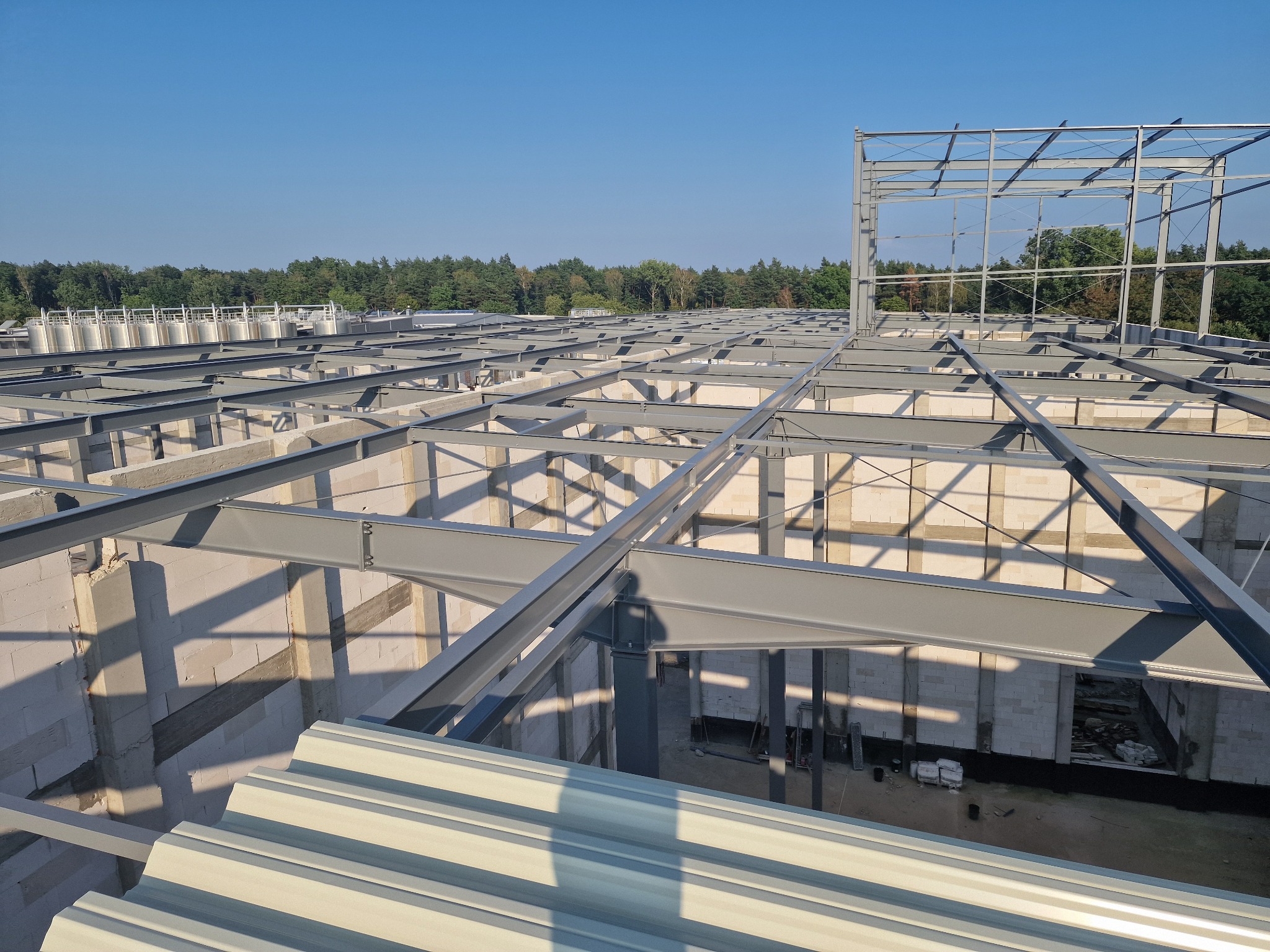 Konstrukcja stalowa hali w trakcie budowy, widok z góry na szkielet z belek i słupów, ściany z bloczków, w tle las i zbiorniki.