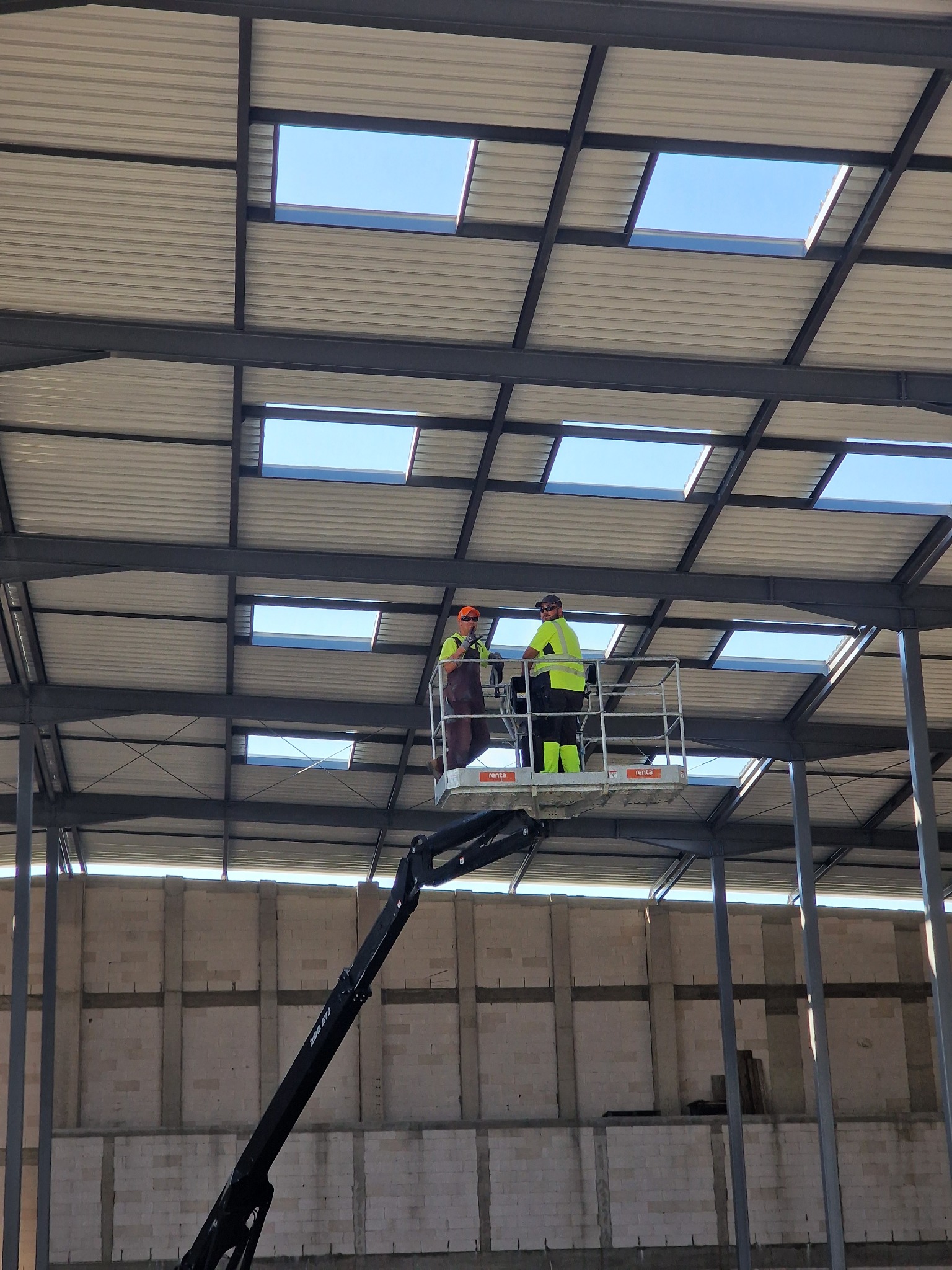 Dwóch pracowników na podnośniku koszowym montuje elementy dachu hali stalowej z świetlikami, widoczne ściany z bloczków betonowych.