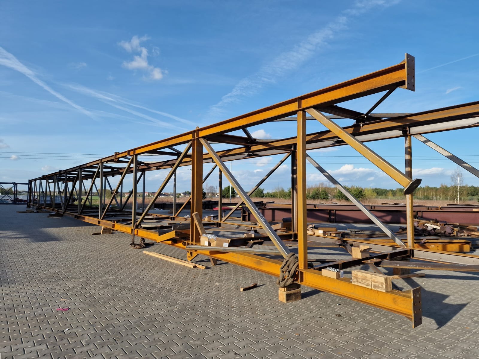 Szkielet stalowej konstrukcji hali w kolorze pomarańczowym, widok z boku na tle błękitnego nieba z wyraźnymi smugami kondensacyjnymi