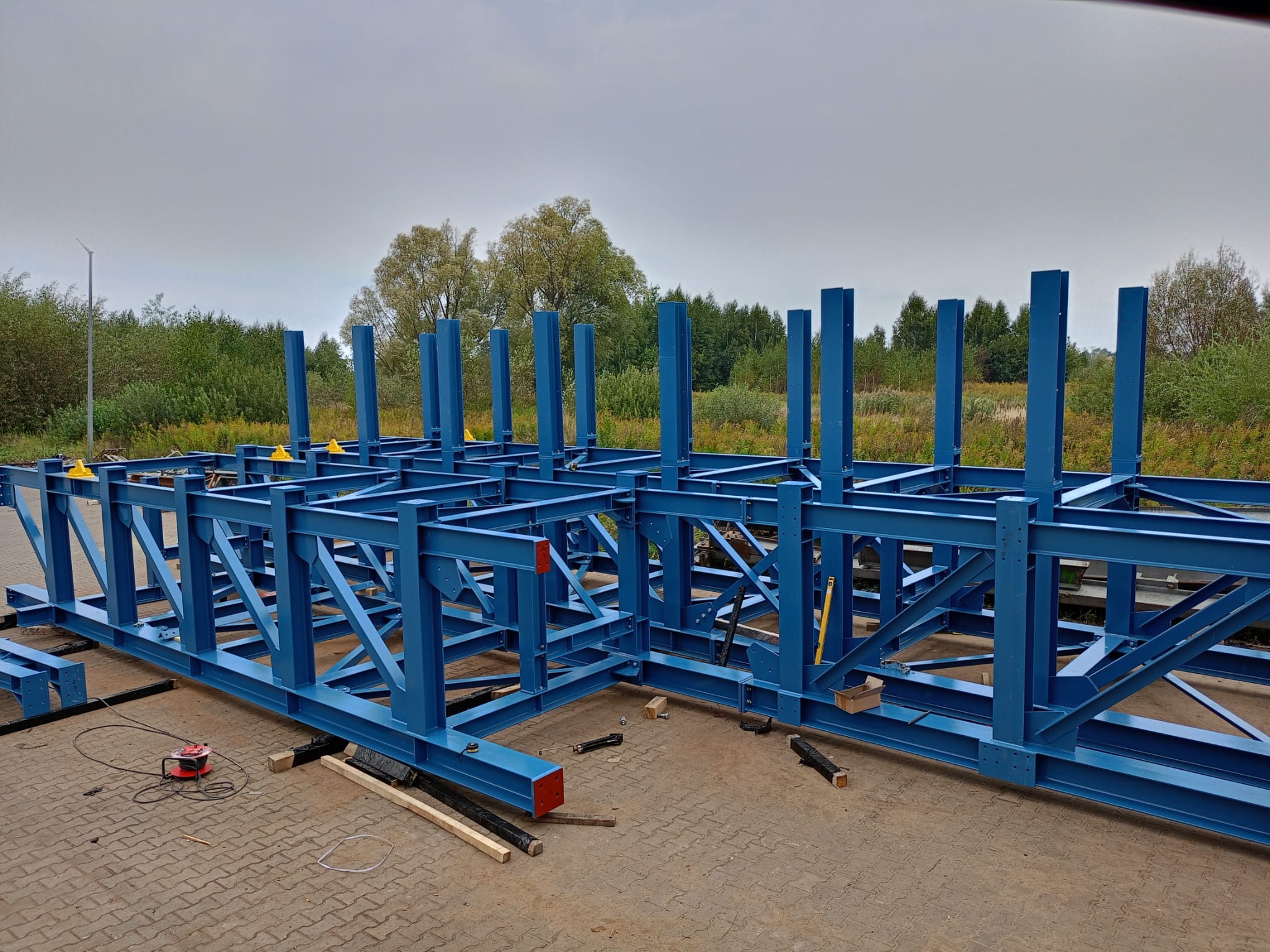 Niebieska, stalowa konstrukcja hali w trakcie montażu, widoczne elementy skręcane i spawane, z żółtymi klinami poziomującymi, na zewnątrz, w tle drzewa.
