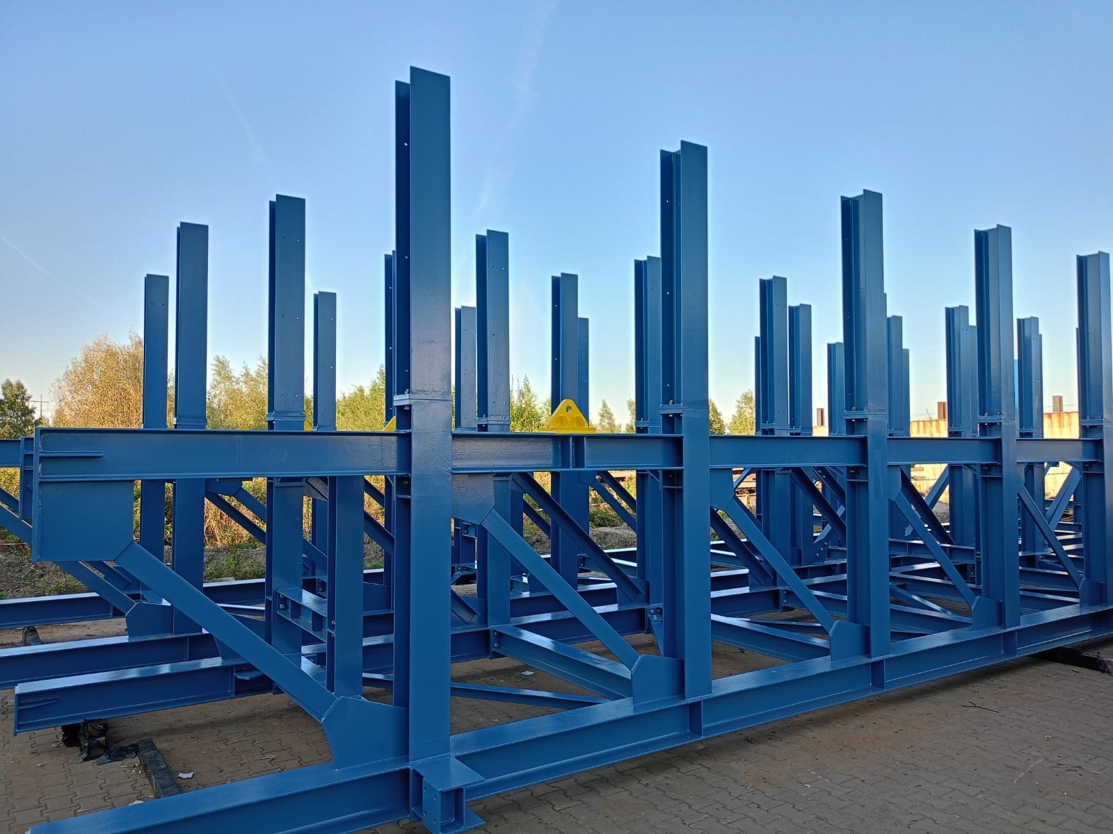 Niebieska konstrukcja stalowa hali w trakcie budowy, widoczne słupy i belki, żółte elementy zabezpieczające, w tle drzewa i błękitne niebo.