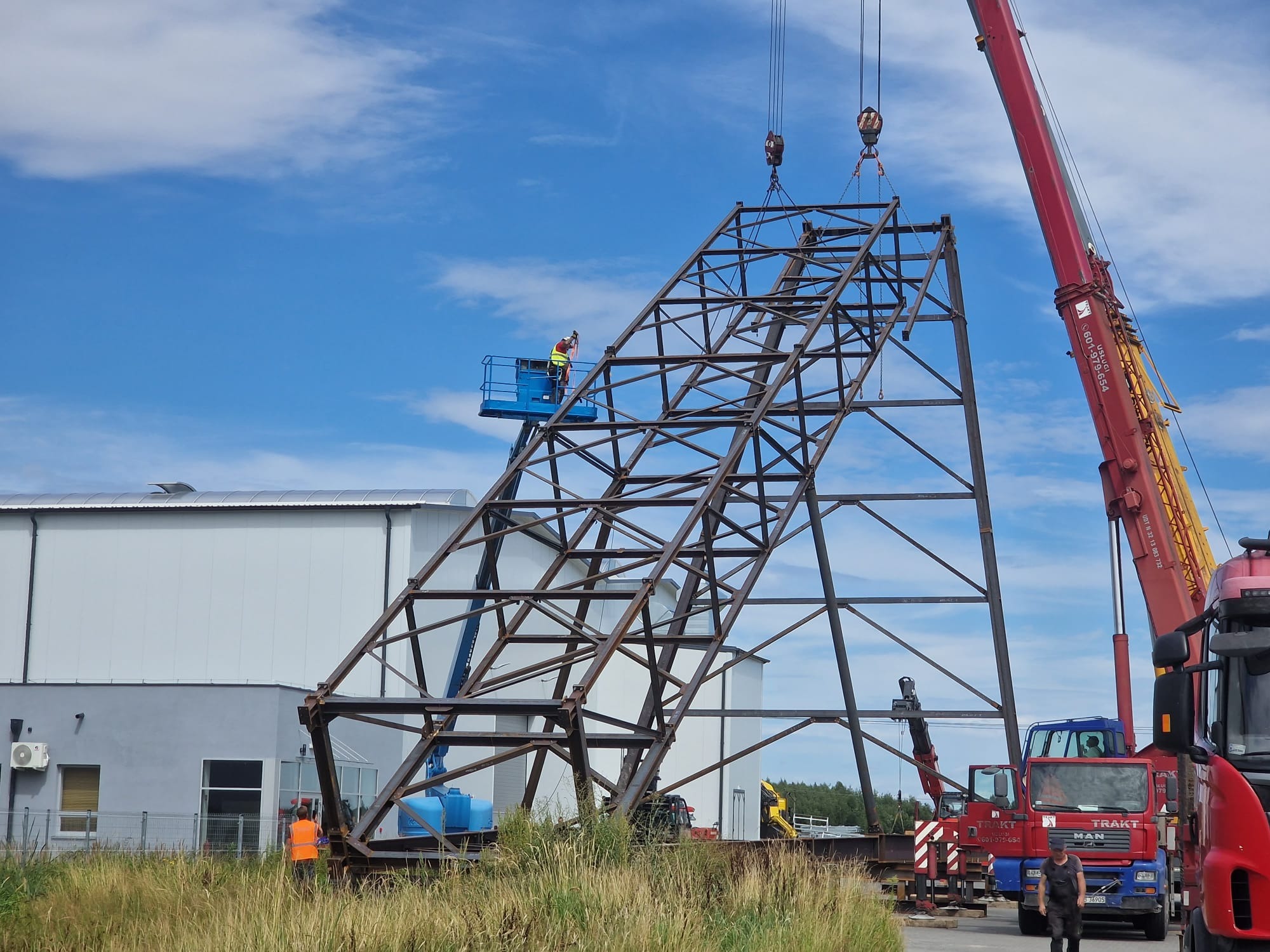 Montaż stalowej konstrukcji kratownicowej za pomocą dźwigu i podnośnika koszowego na tle hali przemysłowej i błękitnego nieba.