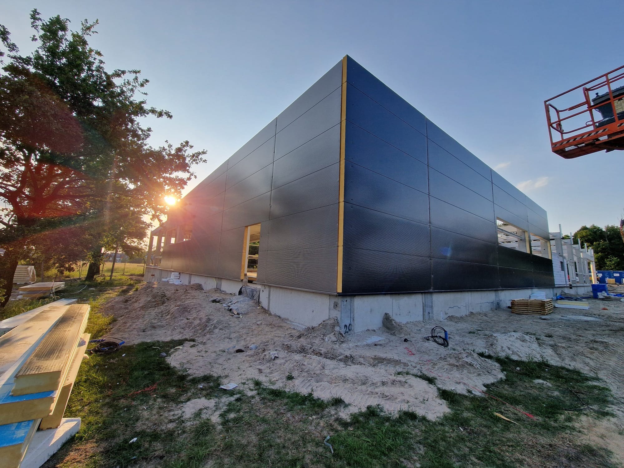Budowa hali: widok na budynek z czarnymi panelami, częściowo ukończony, z widocznymi elementami konstrukcyjnymi i stertą paneli obok, w tle drzewa i fragment podnośnika.