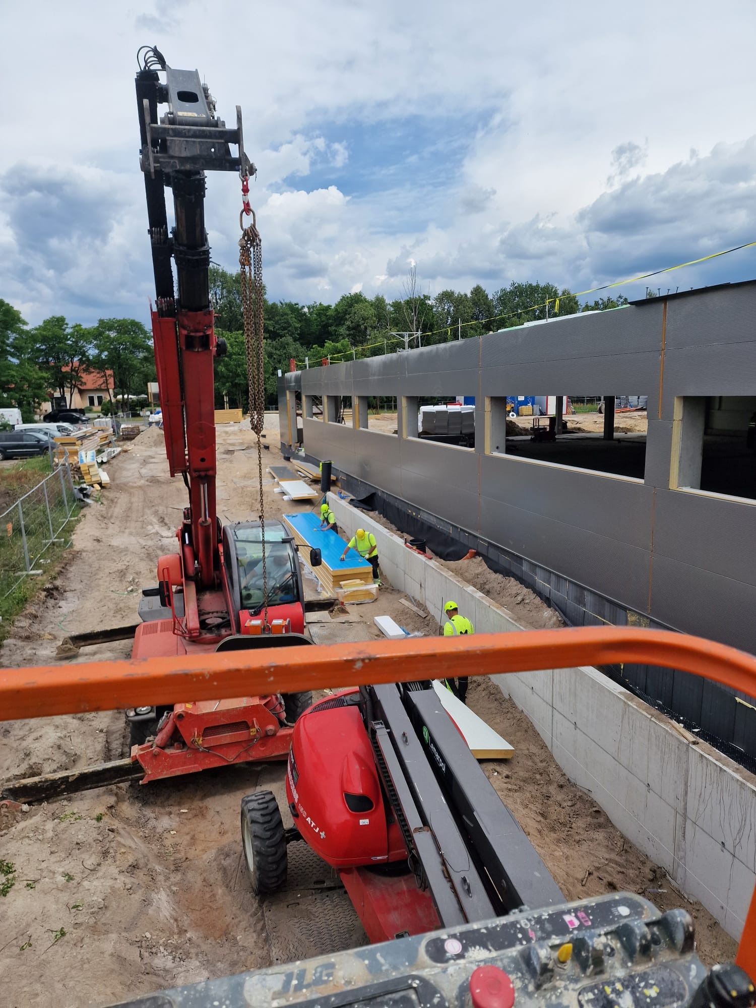 Czerwony dźwig podnosi płyty na placu budowy hali z szarymi ścianami, pracownicy w żółtych kamizelkach przygotowują elementy do montażu.
