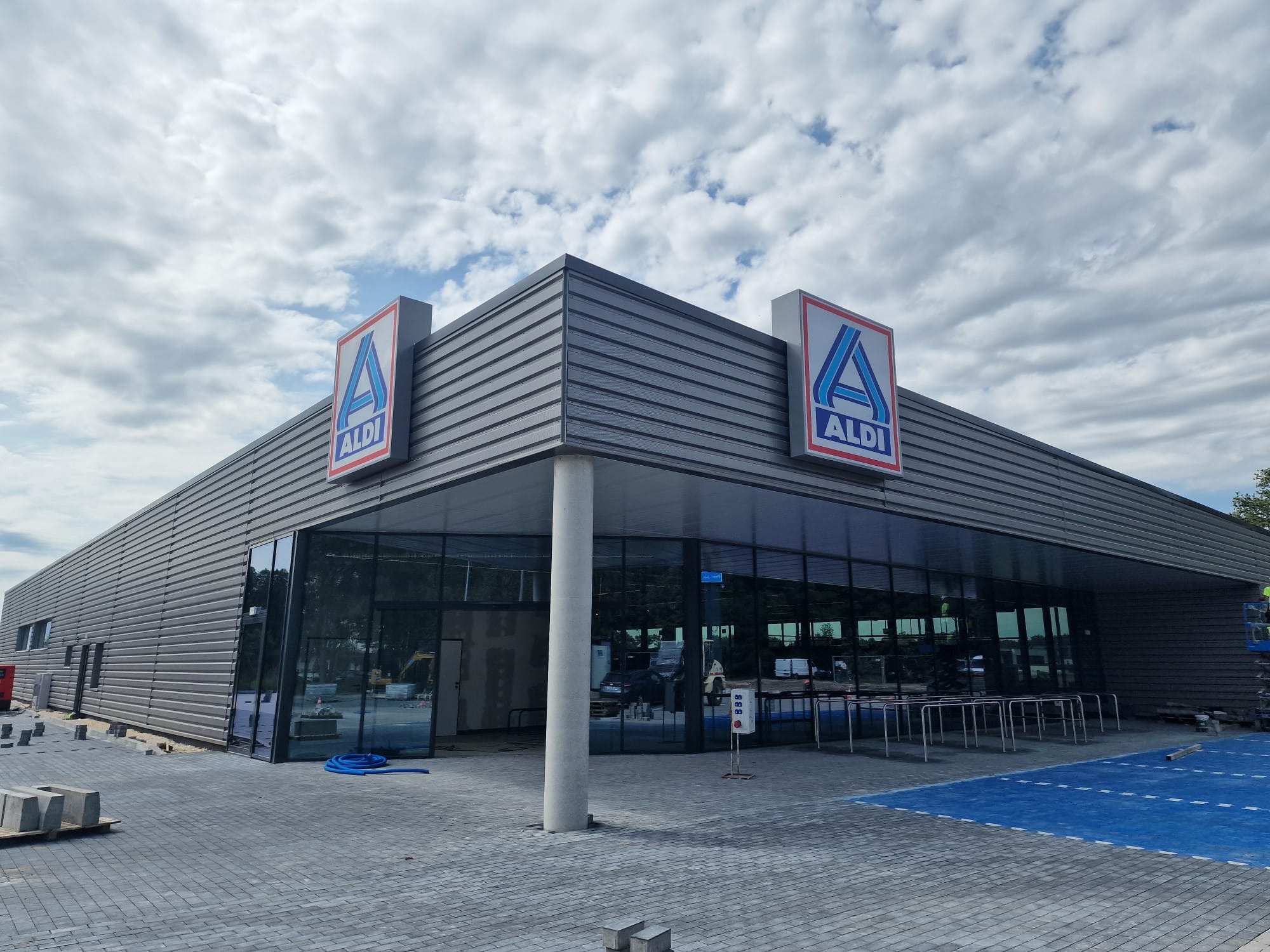 Nowo wybudowany budynek sklepu z szarymi panelami elewacyjnymi, dużymi oknami i logo na elewacji, otoczony kostką brukową i elementami wykończeniowymi.