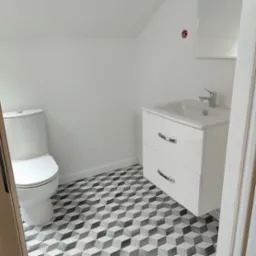 Nowoczesna łazienka w odcieniach bieli z toaletą, szafką umywalkową z dwoma szufladami i geometryczną podłogą w 3D wzór; widoczne niedokończone gniazdko elektryczne.