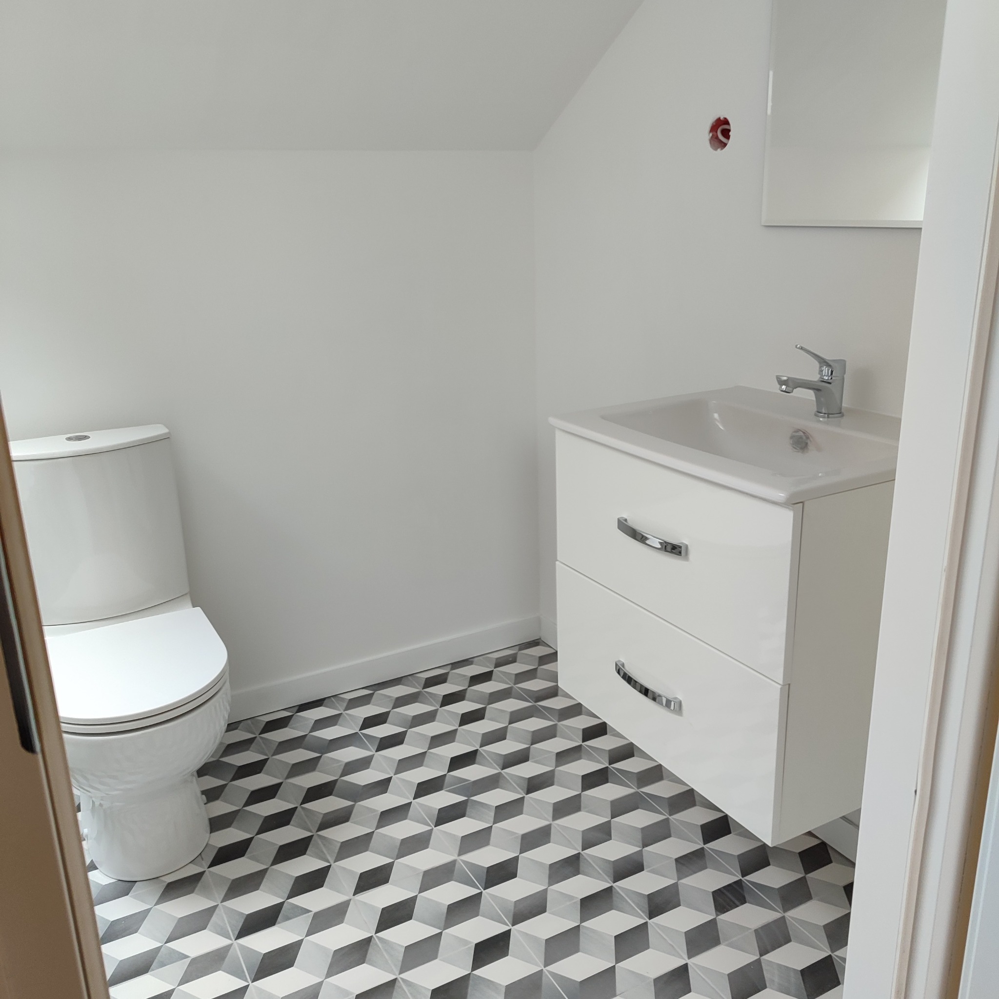 Nowoczesna łazienka w odcieniach bieli z toaletą, szafką umywalkową z dwoma szufladami i geometryczną podłogą w 3D wzór; widoczne niedokończone gniazdko elektryczne.