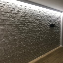 Housesett Tomasz Podkanowicz - Ściana wyłożona szarą cegłą dekoracyjną z podświetleniem LED u góry, widoczny fragment drewnianej podłogi i uchwyt na telewizor.