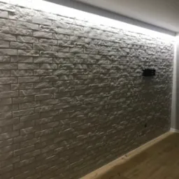 Ściana wyłożona szarą cegłą dekoracyjną z podświetleniem LED u góry, widoczny fragment drewnianej podłogi i uchwyt na telewizor.