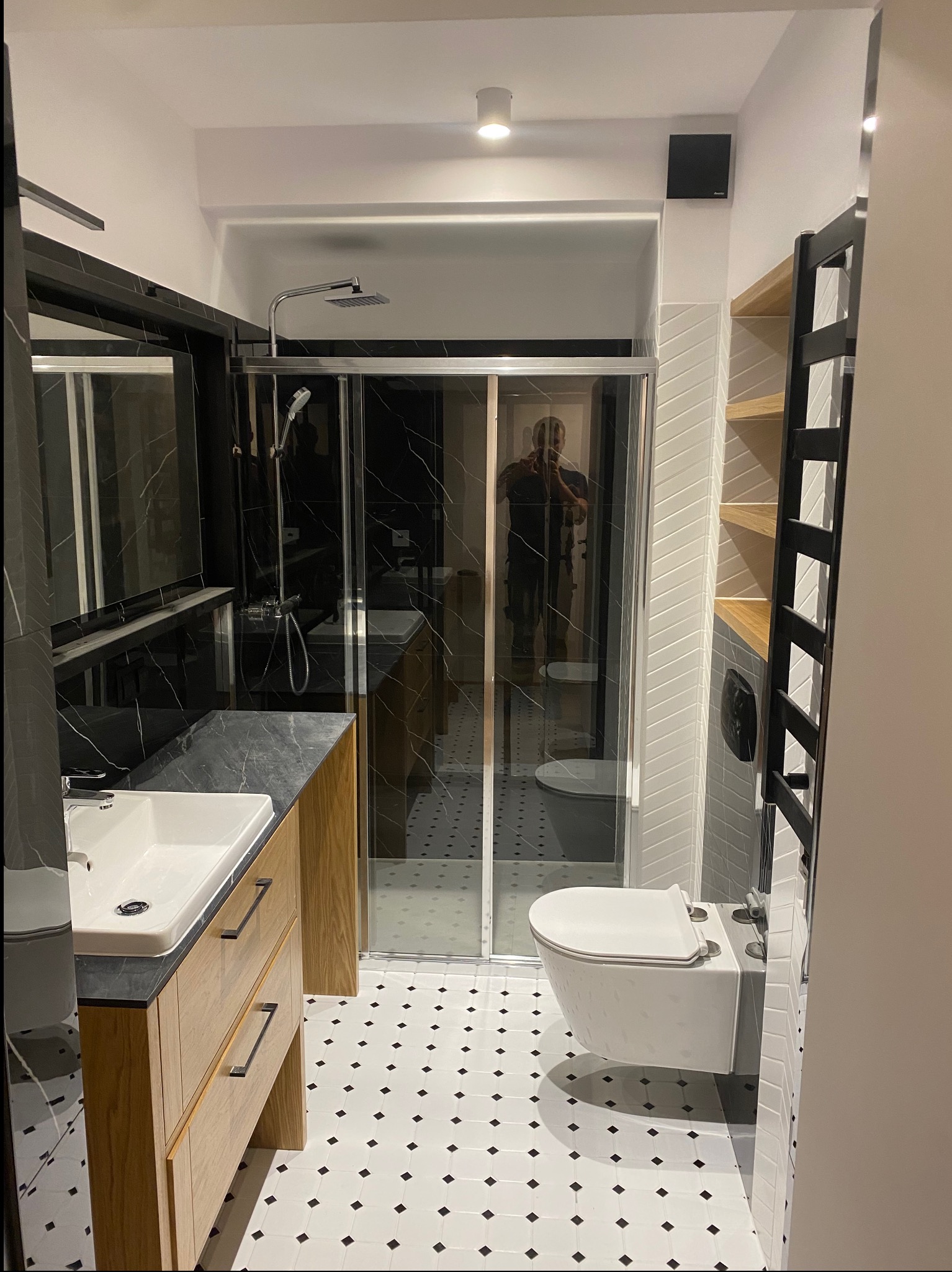 Nowoczesna łazienka po remoncie z kabiną prysznicową z czarnym marmurem, umywalką nablatową na szafce z jasnego drewna, wiszącą toaletą i biało-czarną mozaiką na podłodze.