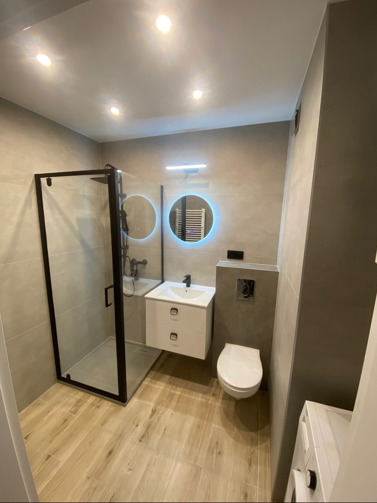 Nowoczesna łazienka z kabiną prysznicową typu walk-in z czarnymi ramami, podświetlanym okrągłym lustrem i drewnopodobną podłogą, widok na toaletę i pralkę.