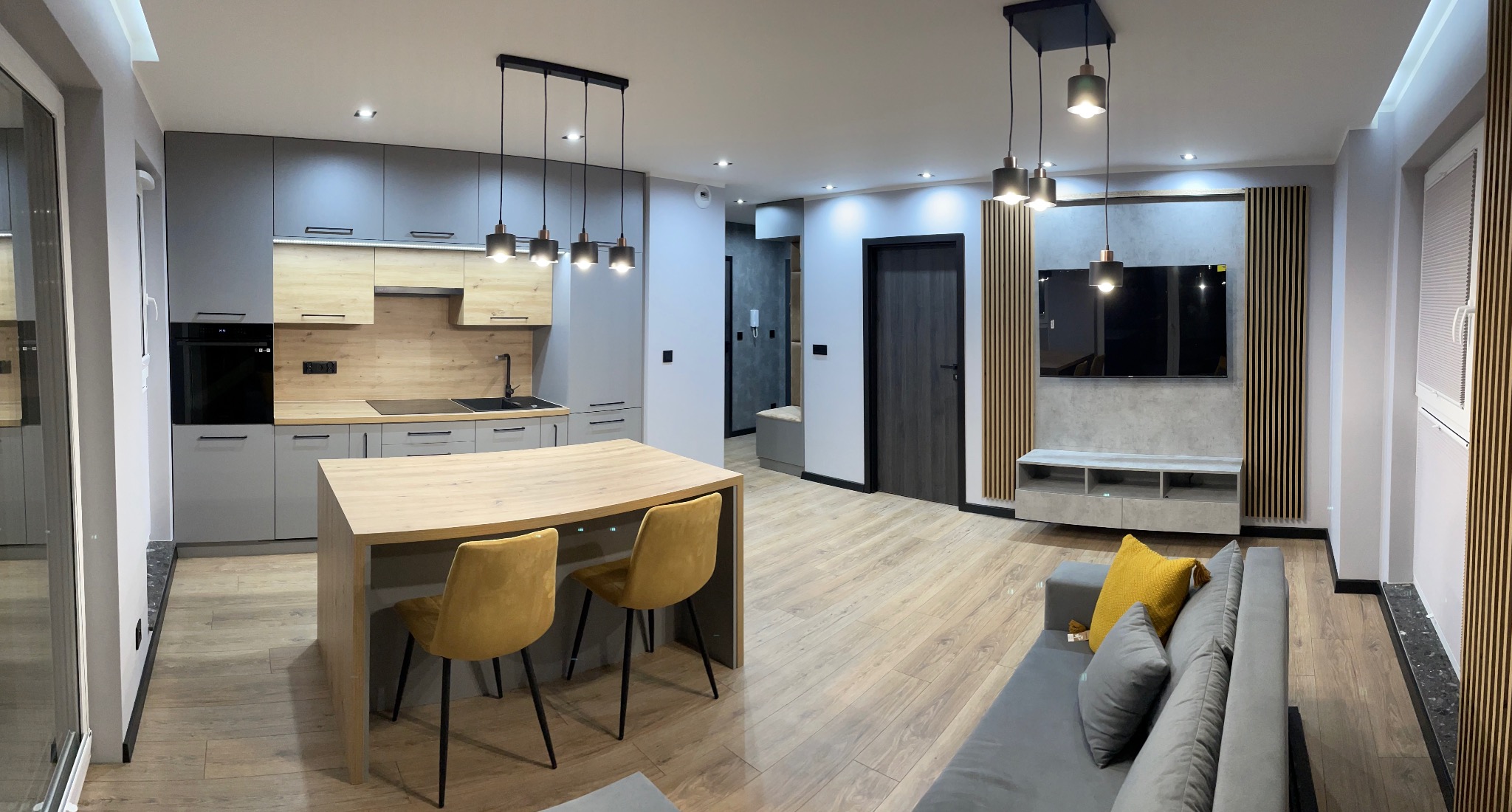 Panoramiczne ujęcie nowoczesnego wnętrza: szara zabudowa kuchenna z drewnianymi akcentami, wyspa z krzesłami w kolorze musztardowym, drewniana podłoga, ściana telewizyjna z dekoracyjnymi panelami...