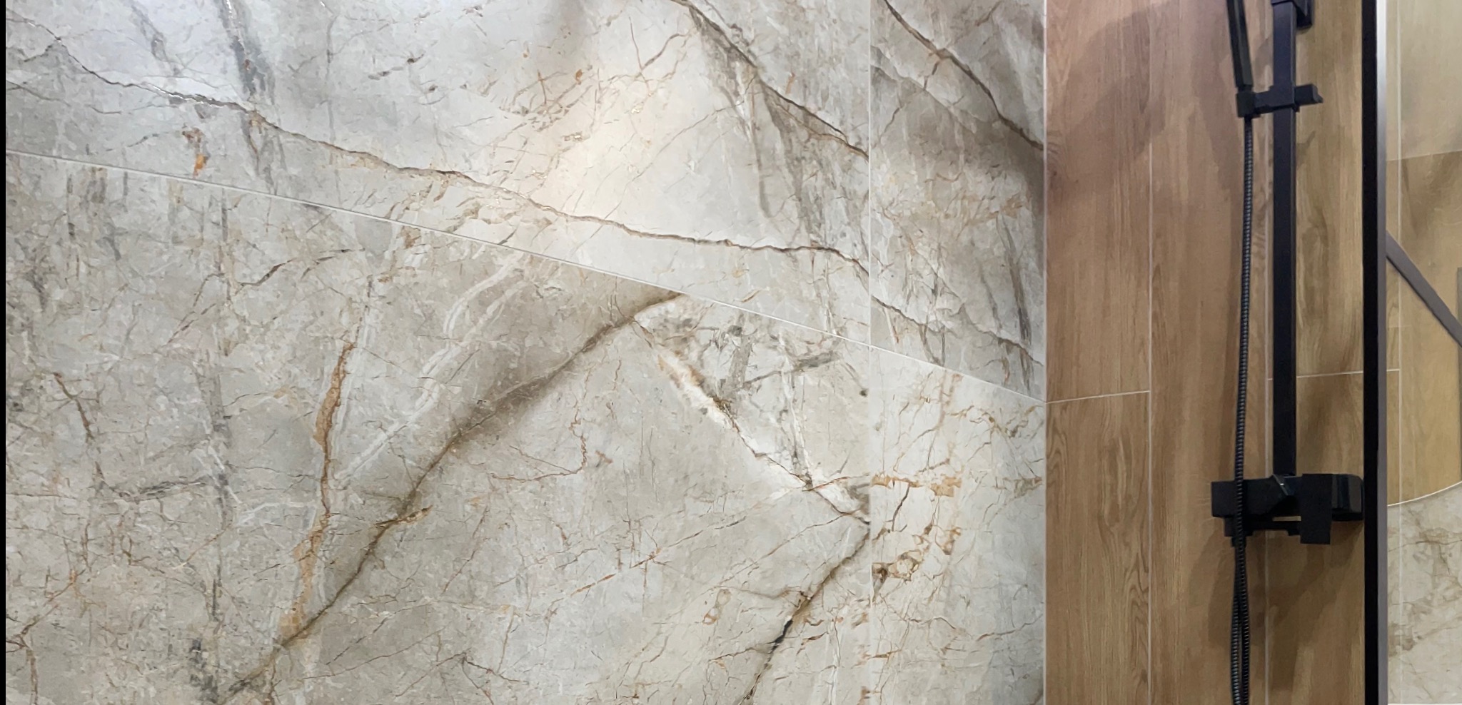 Beżowo-szara ściana łazienki wyłożona płytkami imitującymi marmur z widoczną czarną armaturą prysznicową.