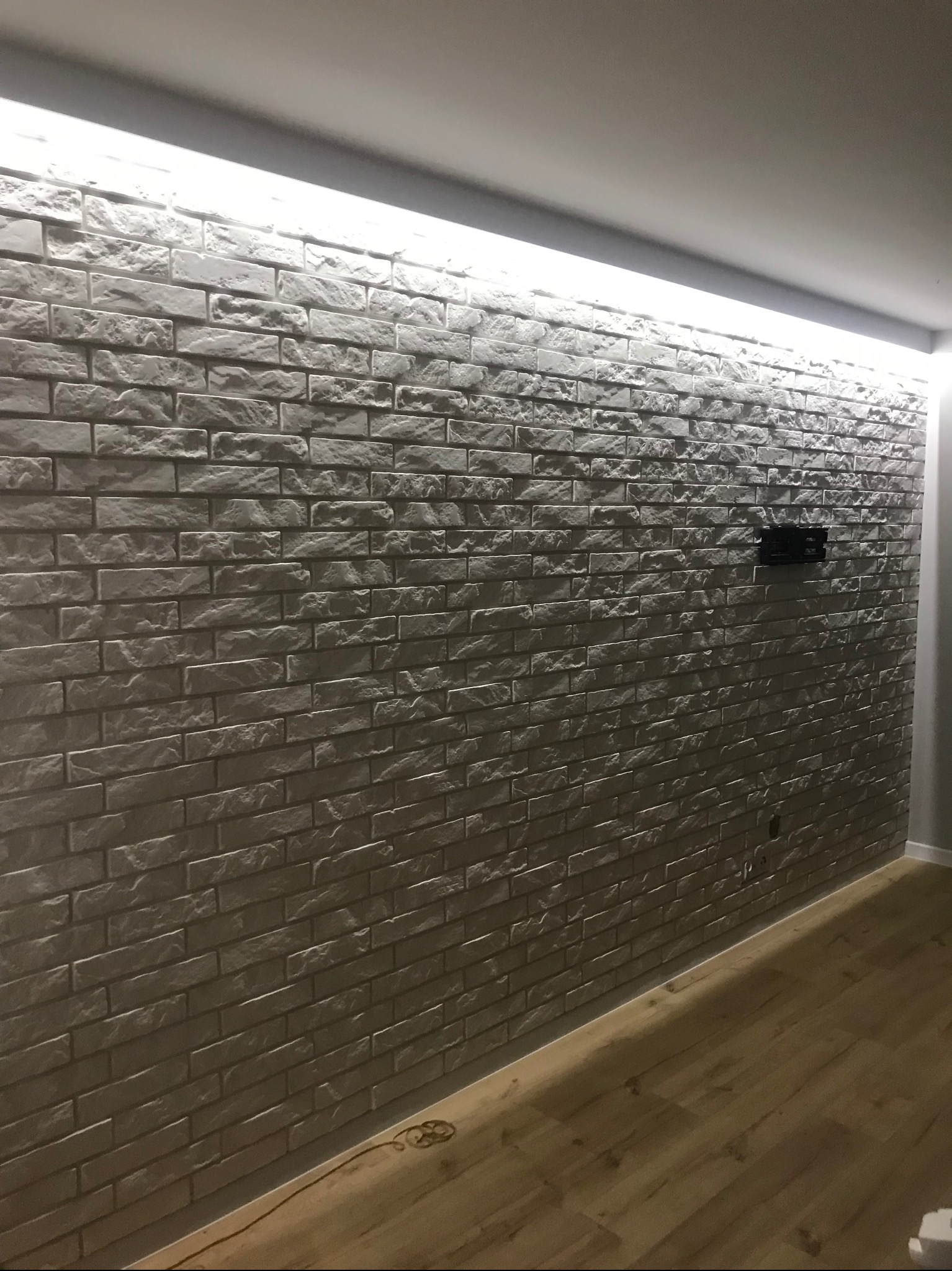Ściana wyłożona szarą cegłą dekoracyjną z podświetleniem LED u góry, widoczny fragment drewnianej podłogi i uchwyt na telewizor.