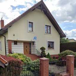 Alpers Mycie Malowanie dachów i elewacji - Dwupiętrowy dom z jasną elewacją, brązowym dachem i stolarką okienną, widoczny zza ozdobnego, niskiego ogrodzenia i zadbanego ogrodu z krzewami.