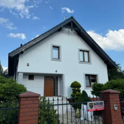 Biały dom jednorodzinny z ciemnym dachem, widoczny od frontu, z ogrodzeniem i tabliczką Solid Security, błękitne niebo z chmurami w tle.