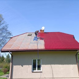 Alpers Mycie Malowanie dachów i elewacji - Częściowo pomalowany dach z dachówki ceramicznej na czerwono, widoczny dekarz podczas pracy, czysty błękit nieba w tle.