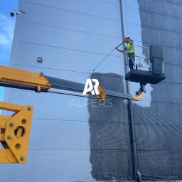 Alpers Mycie Malowanie dachów i elewacji - Pracownik w żółtym kasku i kamizelce, na podnośniku koszowym, czyści elewację hali przemysłowej w Starych Babicach za pomocą myjki ciśnieniowej. Widoczny kontrast między umytą a brudną powierzchnią.