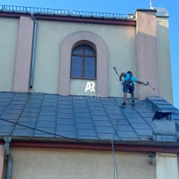 Alpers Mycie Malowanie dachów i elewacji - Pracownik w uprzęży alpinistycznej na dachu budynku sakralnego, prawdopodobnie podczas prac konserwacyjnych, z widocznym kominem i rynnami.
