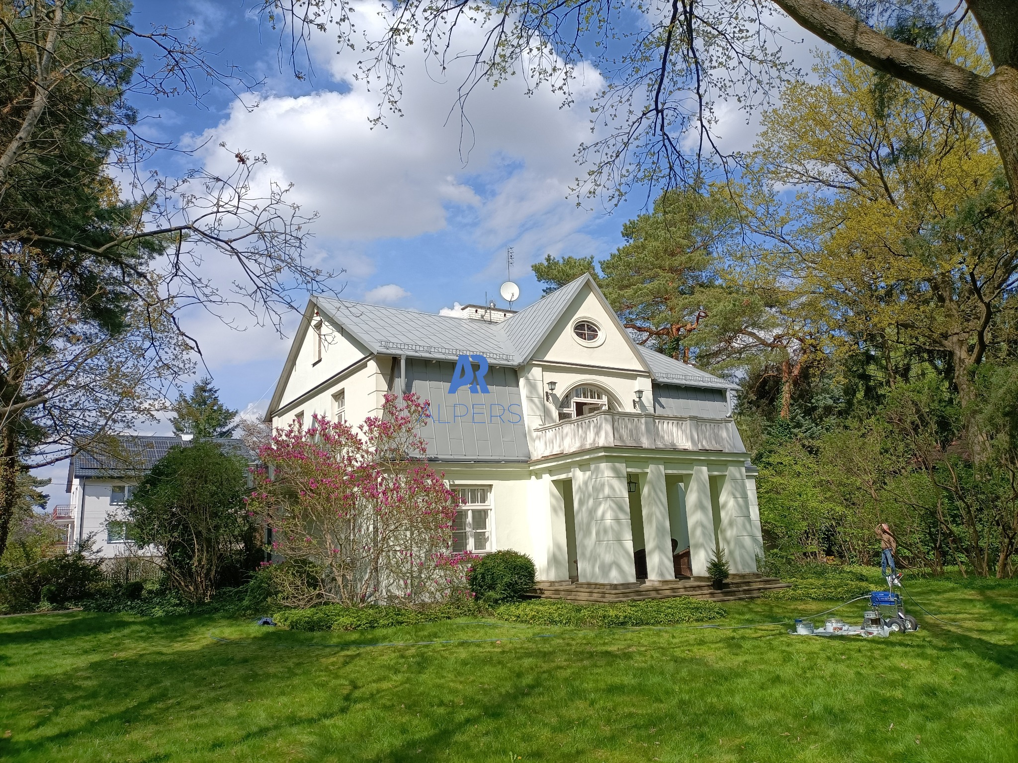 Malowanie elewacji białego domu z kolumnami; widoczny agregat malarski i pracownik w oddali, zielony trawnik i kwitnący krzew magnolii na pierwszym planie.