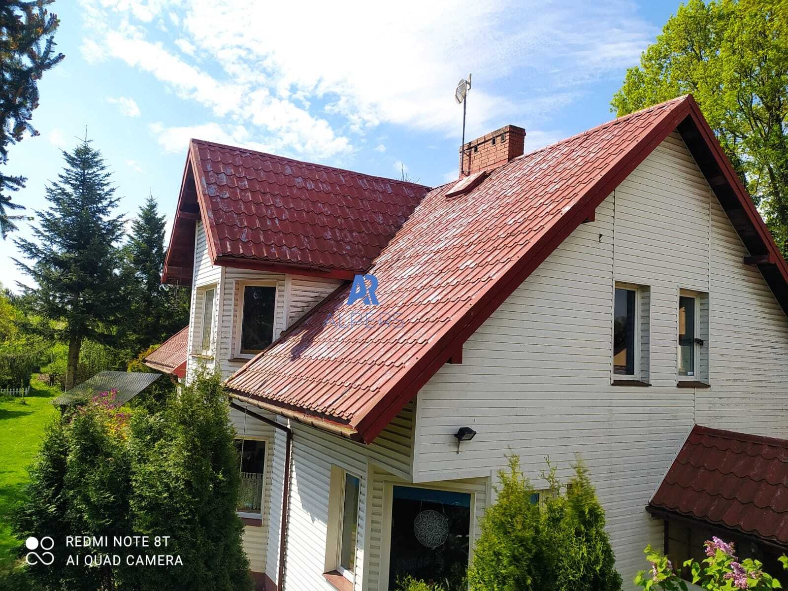 Dwupiętrowy dom z białymi ścianami i czerwoną dachówką, widoczny komin z anteną, otoczony zielenią, na dachu miejscami widoczne zabrudzenia.