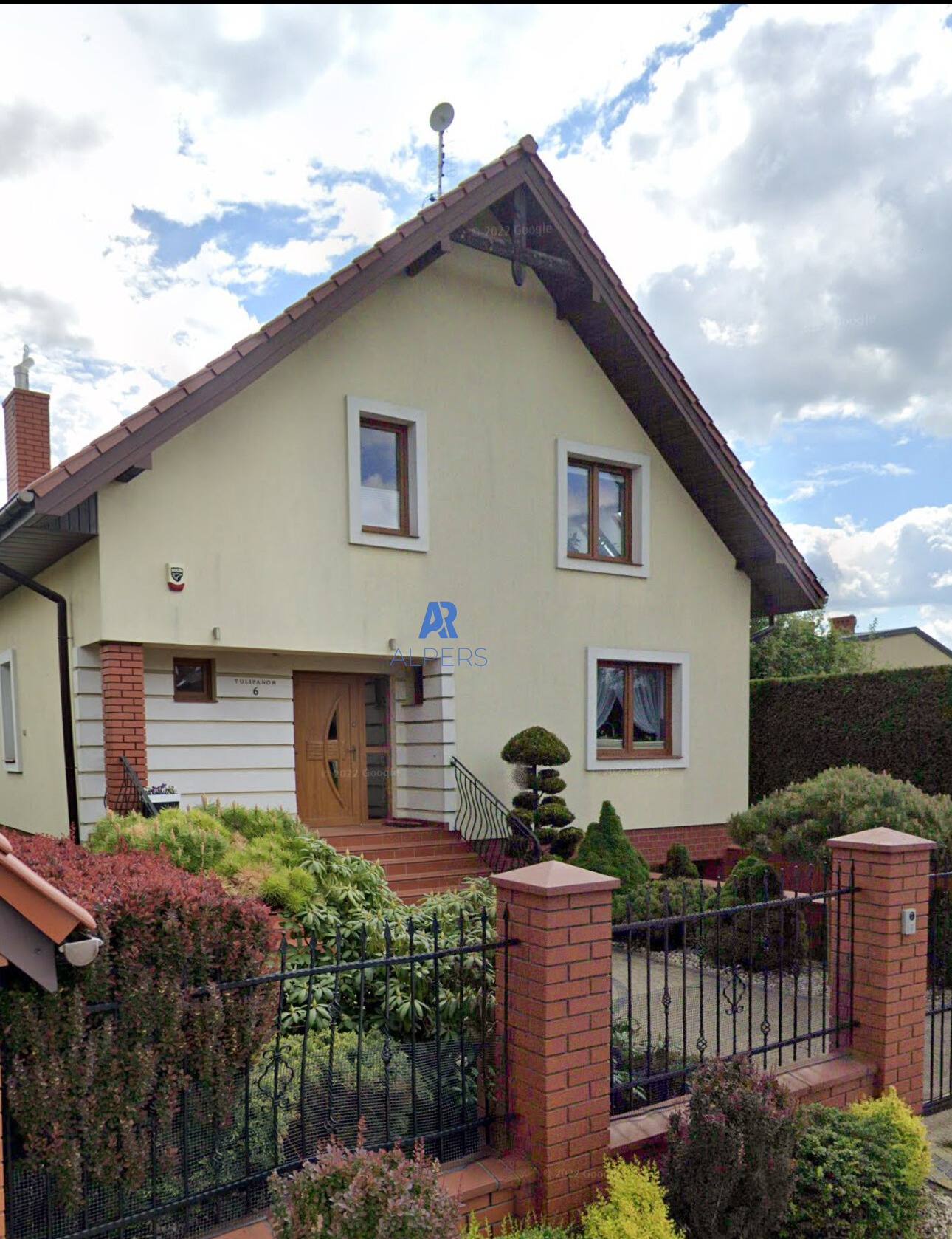 Dwupiętrowy dom z jasną elewacją, brązowym dachem i stolarką okienną, widoczny zza ozdobnego, niskiego ogrodzenia i zadbanego ogrodu z krzewami.