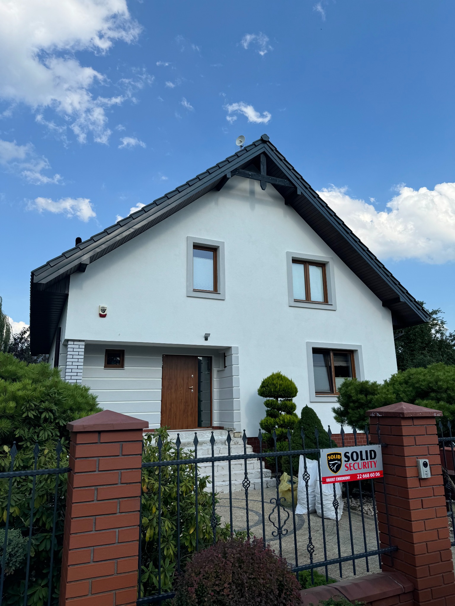Biały dom jednorodzinny z ciemnym dachem, widoczny od frontu, z ogrodzeniem i tabliczką Solid Security, błękitne niebo z chmurami w tle.