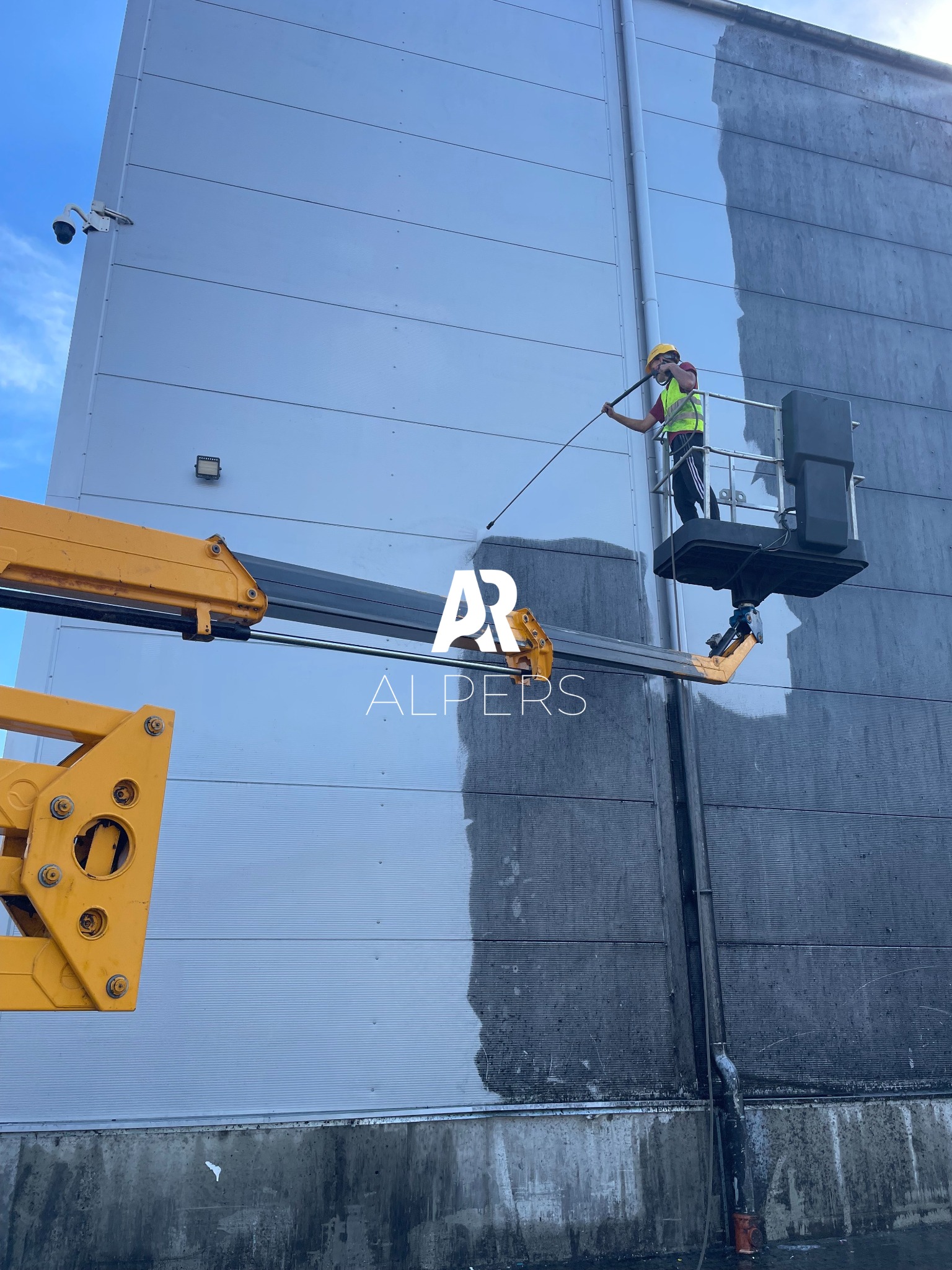 Pracownik w żółtym kasku i kamizelce, na podnośniku koszowym, czyści elewację hali przemysłowej w Starych Babicach za pomocą myjki ciśnieniowej. Widoczny kontrast między umytą a brudną powierzchnią.