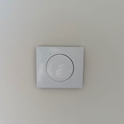 WOLWO Wojciech Wolski - Biały, kwadratowy włącznik światła z okrągłym przyciskiem, zamontowany na jasnej, gładkiej ścianie. Minimalistyczny design, centralne ujęcie.