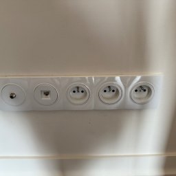 WOLWO Wojciech Wolski - Biała listwa z gniazdem antenowym, gniazdem RJ45 oraz trzema gniazdami elektrycznymi, zamontowana na białej ścianie, widok od dołu.