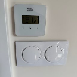 WOLWO Wojciech Wolski - Biały, kwadratowy termostat TECH Sterowniki z wyświetlaną temperaturą 21.2 stopni Celsjusza, zamontowany nad podwójnym, okrągłym włącznikiem światła na kremowej ścianie.