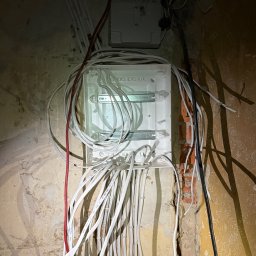 WOLWO Wojciech Wolski - Biała skrzynka elektryczna z licznymi kablami wchodzącymi i wychodzącymi, zamontowana na surowej ścianie z fragmentami odsłoniętej cegły i starymi tapetami.