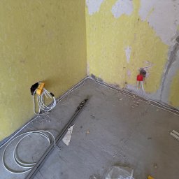 WOLWO Wojciech Wolski - Wnętrze w trakcie remontu, widoczne fragmenty zerwanej żółtej tapety, kable elektryczne prowadzone po ścianie i podłodze, narzędzie elektryczne z nawiniętym kablem przy ścianie, oraz listwy...