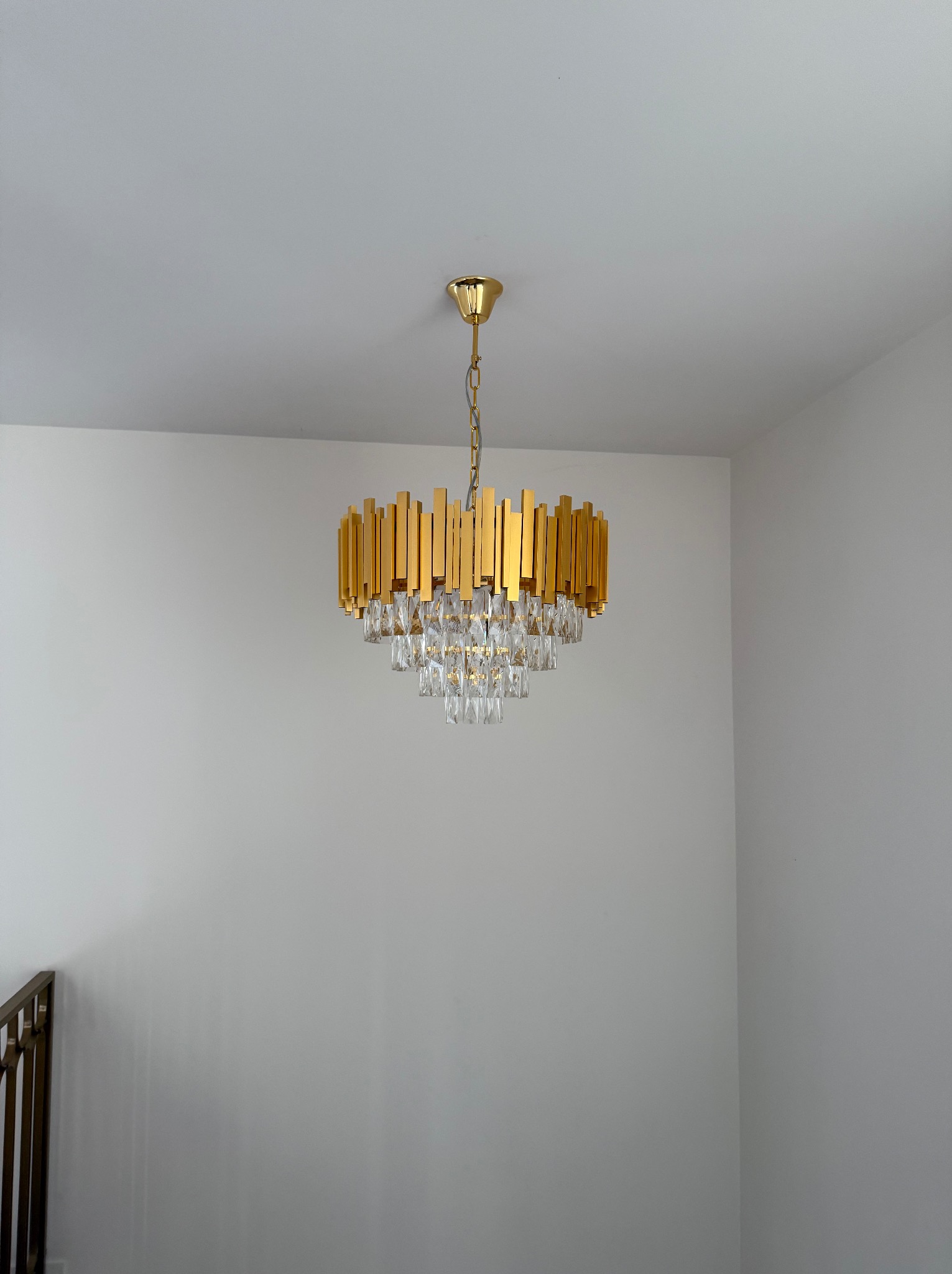 Złoty żyrandol z kryształkami, zamontowany na białym suficie w rogu pomieszczenia, widoczny fragment metalowej balustrady.