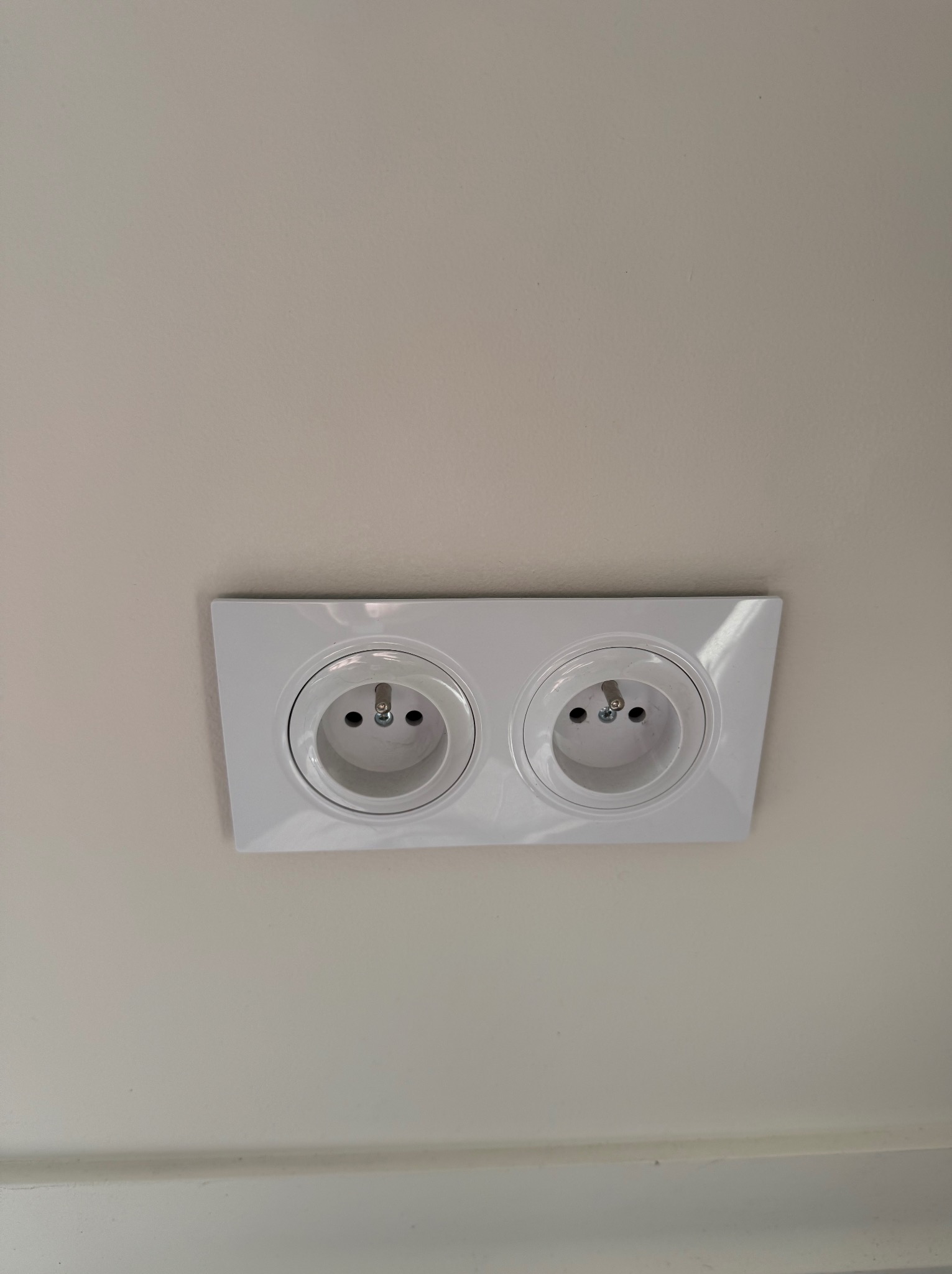 Białe, podwójne gniazdko elektryczne zamontowane na jasnej ścianie, widok z dołu.