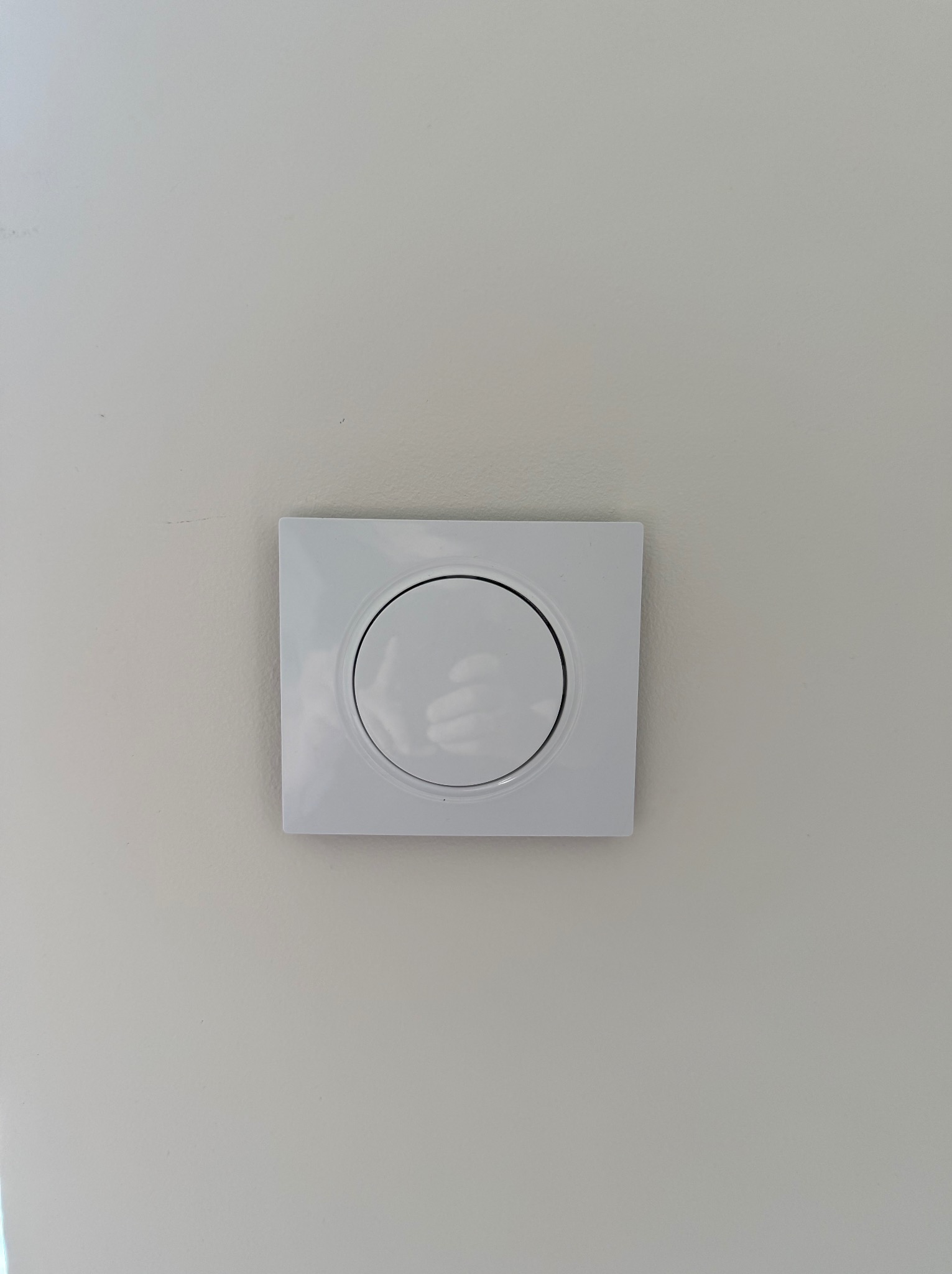 Biały, kwadratowy włącznik światła z okrągłym przyciskiem, zamontowany na jasnej, gładkiej ścianie. Minimalistyczny design, centralne ujęcie.