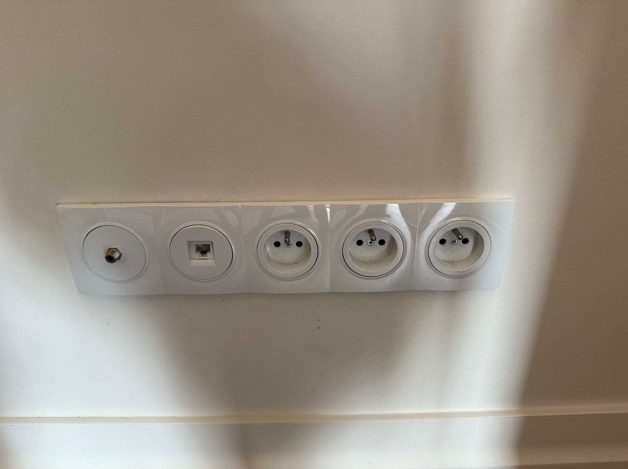 Biała listwa z gniazdem antenowym, gniazdem RJ45 oraz trzema gniazdami elektrycznymi, zamontowana na białej ścianie, widok od dołu.