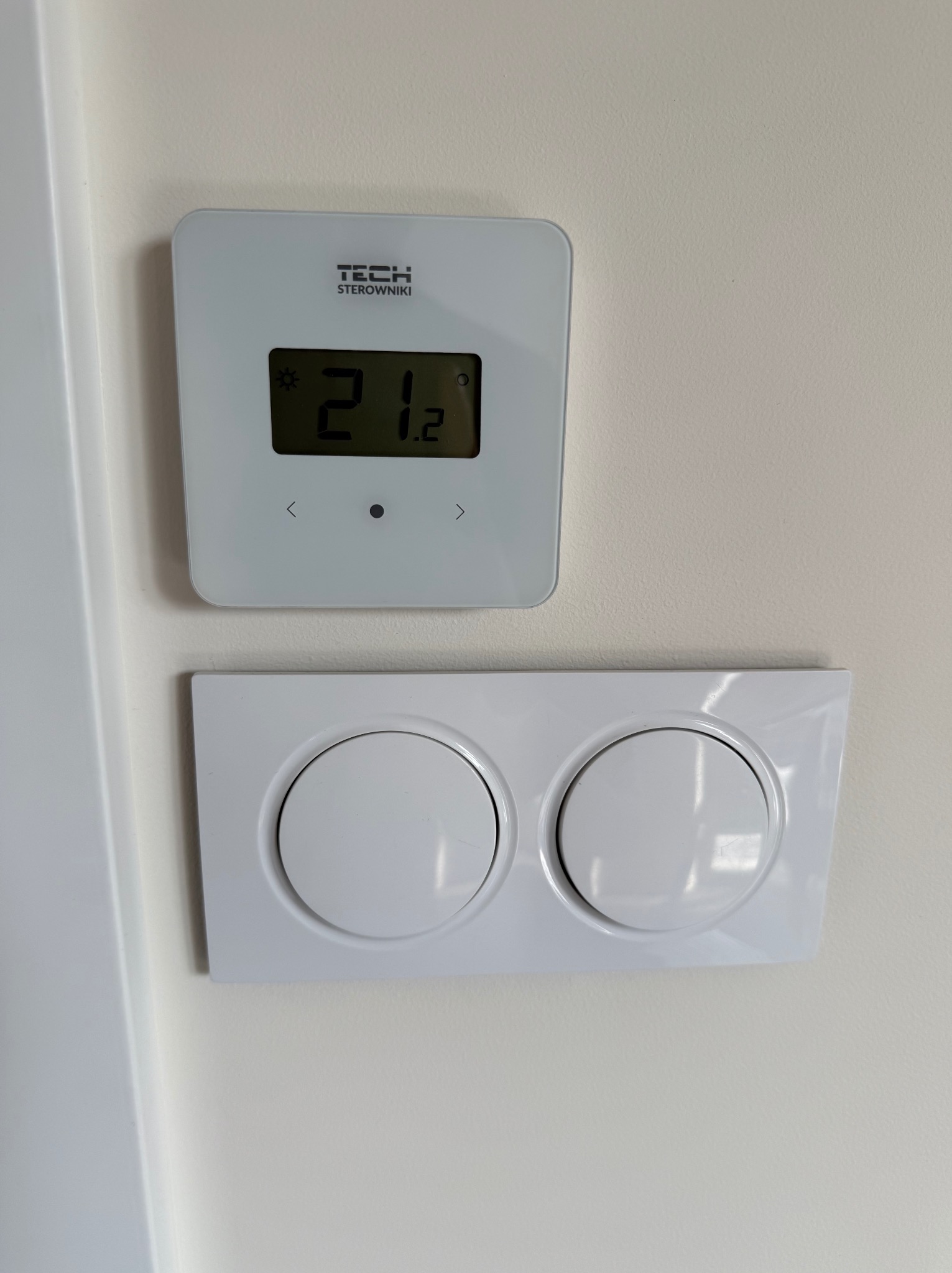 Biały, kwadratowy termostat TECH Sterowniki z wyświetlaną temperaturą 21.2 stopni Celsjusza, zamontowany nad podwójnym, okrągłym włącznikiem światła na kremowej ścianie.