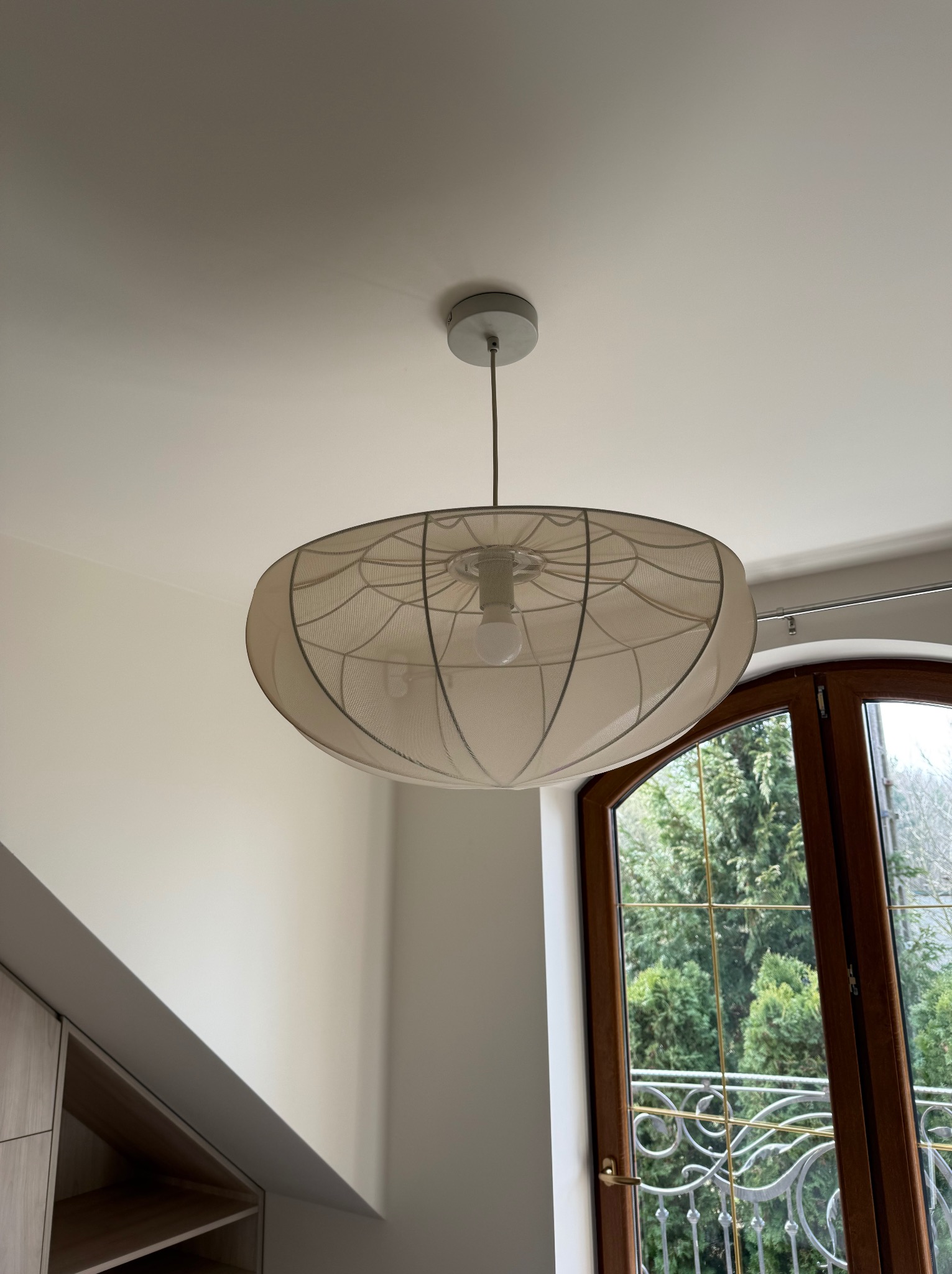 Nowoczesna lampa sufitowa z abażurem w kształcie kuli, zawieszona w jasnym wnętrzu z oknem na ogród.