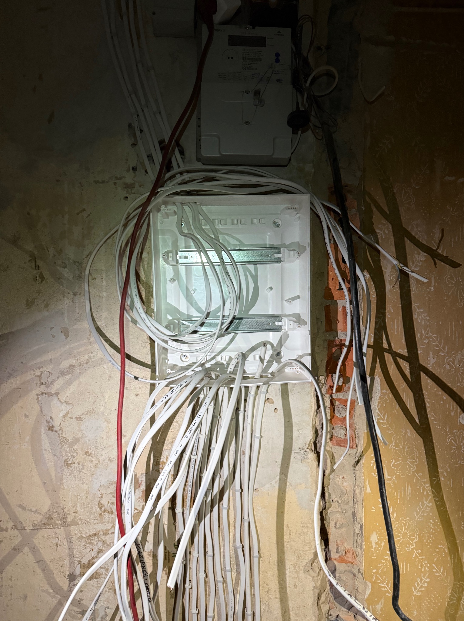 Biała skrzynka elektryczna z licznymi kablami wchodzącymi i wychodzącymi, zamontowana na surowej ścianie z fragmentami odsłoniętej cegły i starymi tapetami.