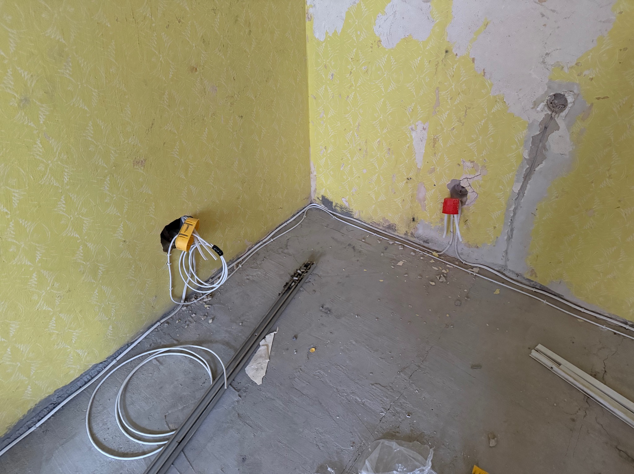 Wnętrze w trakcie remontu, widoczne fragmenty zerwanej żółtej tapety, kable elektryczne prowadzone po ścianie i podłodze, narzędzie elektryczne z nawiniętym kablem przy ścianie, oraz listwy...