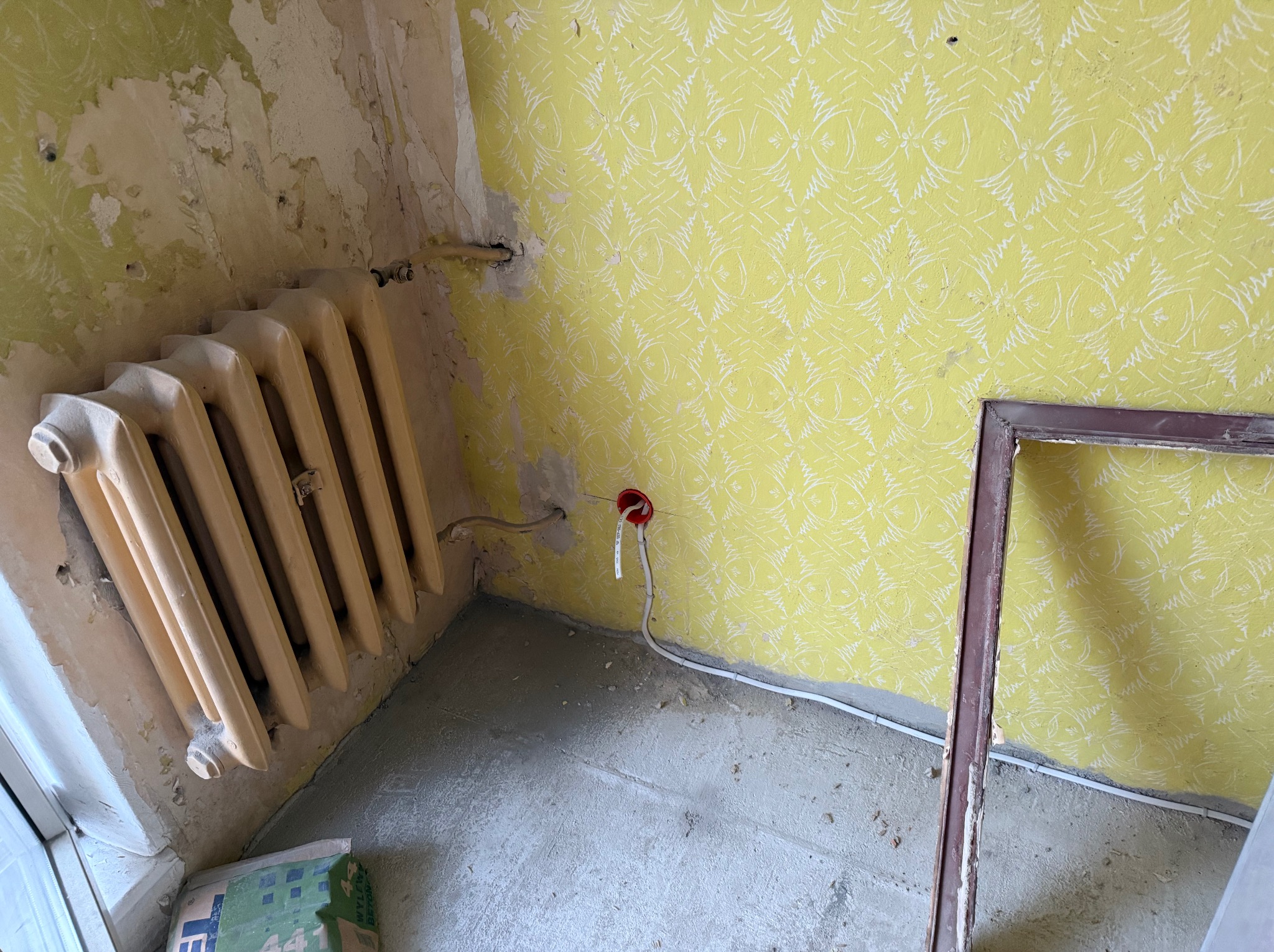 Wnętrze w trakcie remontu: odsłonięta instalacja elektryczna w czerwonej puszce na tle żółtej ściany z wzorzystą tapetą, obok stary grzejnik i rama okienna.