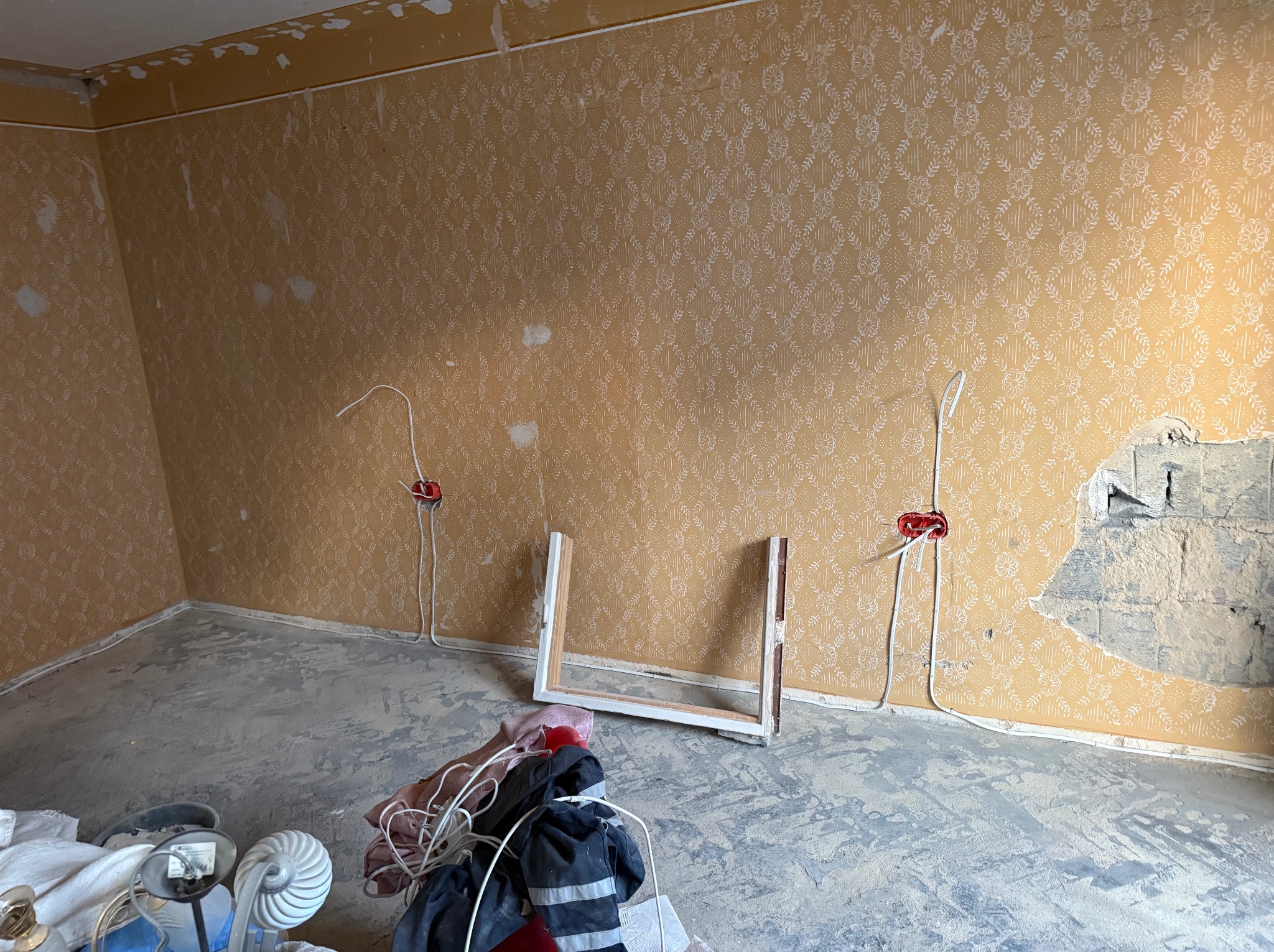 Pokój w trakcie remontu z odsłoniętymi przewodami elektrycznymi i fragmentem ściany bez tapety, widoczne puszki elektryczne z wystającymi kablami, podłoga pokryta warstwą kurzu i resztek materiałów...